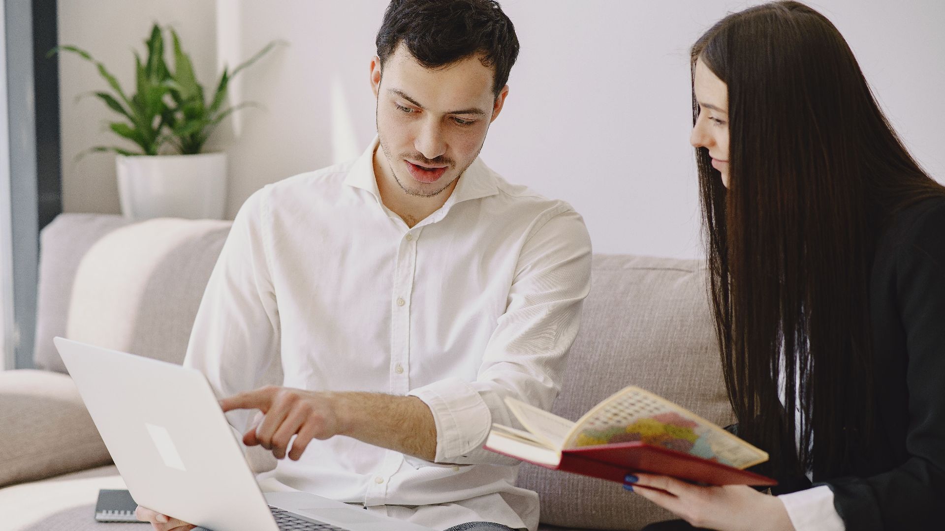 Male worker in formal wear showing with finger at laptop while discussing project with female partner with open copybook and sitting on sofa