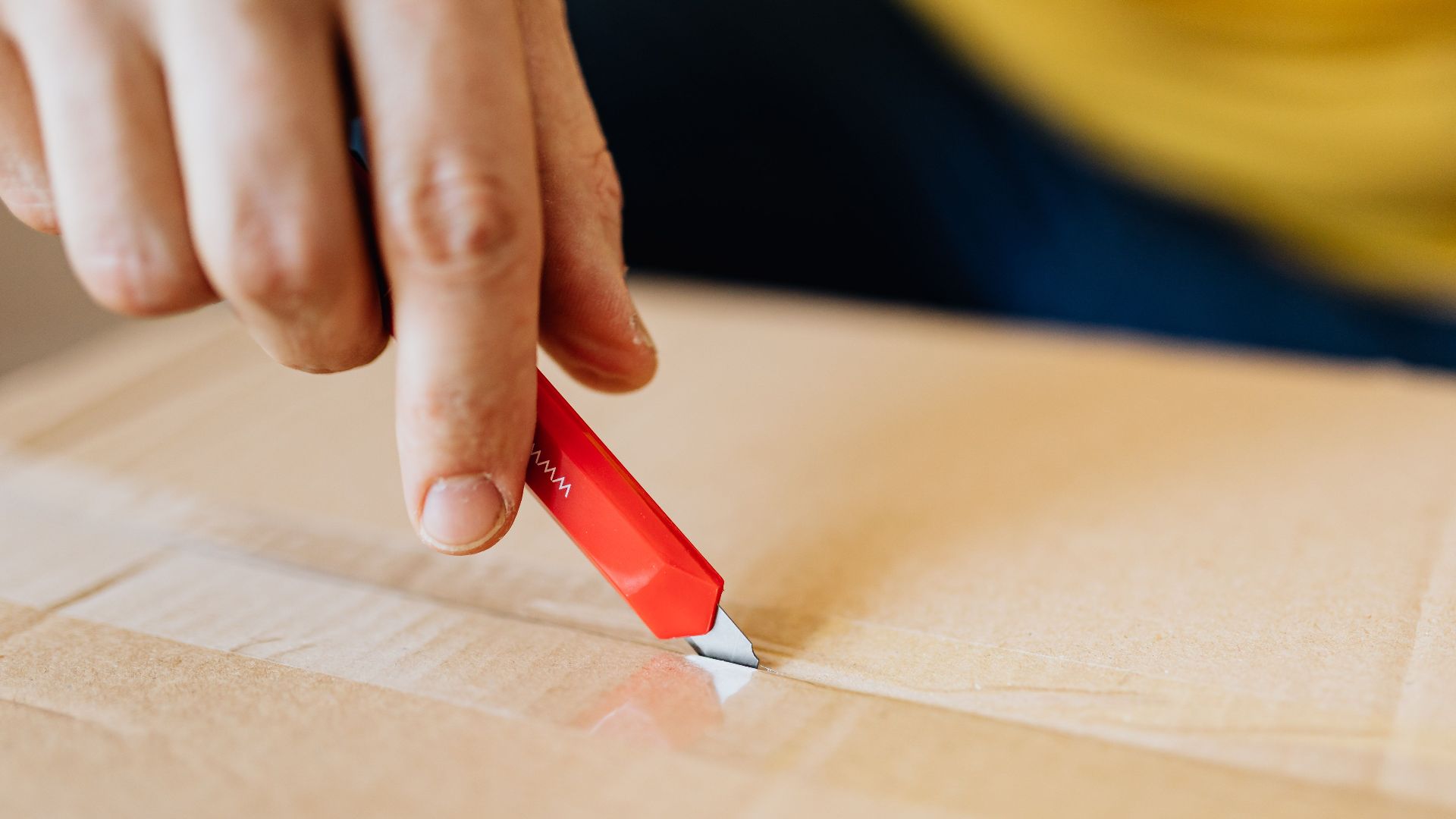 Crop anonymous male unpacking belongings from cardboard box by using sharp utility knife after getting delivery during relocation