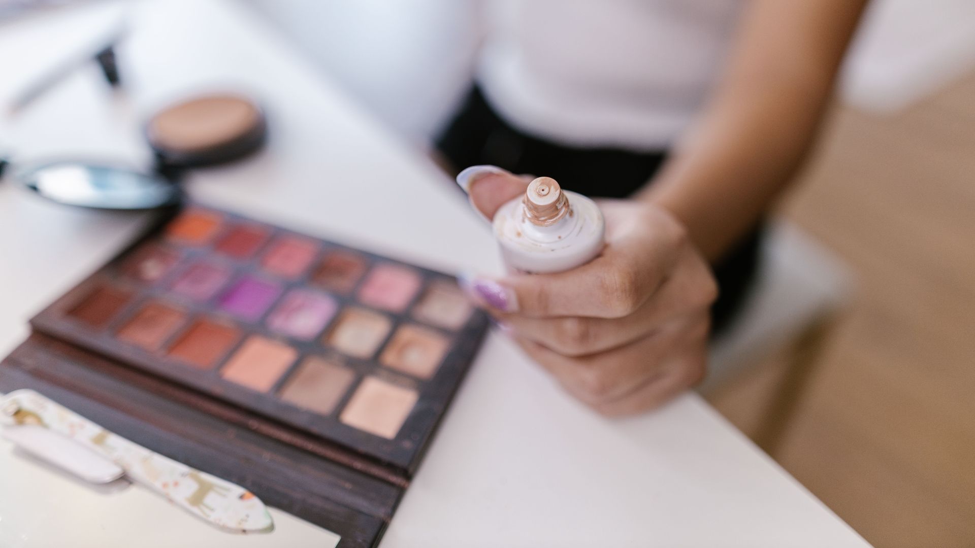 Close-up of a person holding foundation bottle with colorful makeup palette in focus.