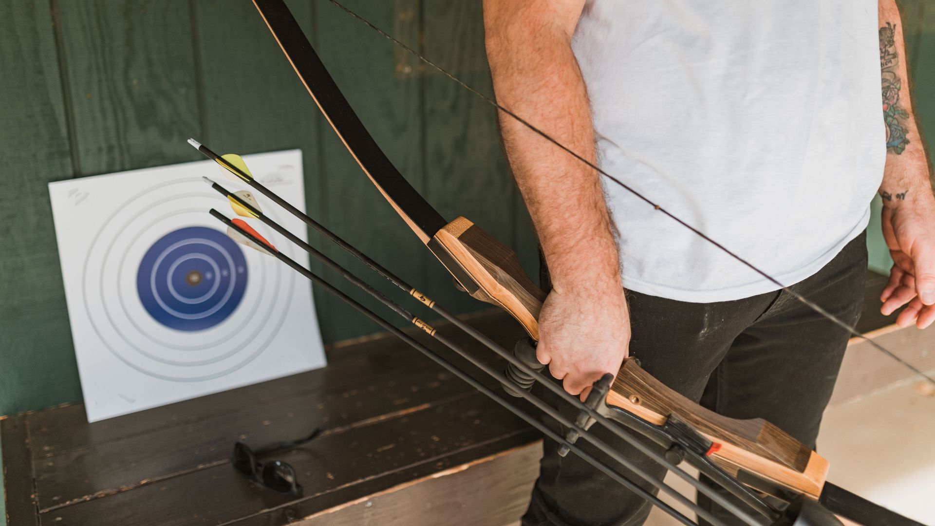 An archer holding a recurve bow with arrows next to a target, in a sunny outdoor setting.