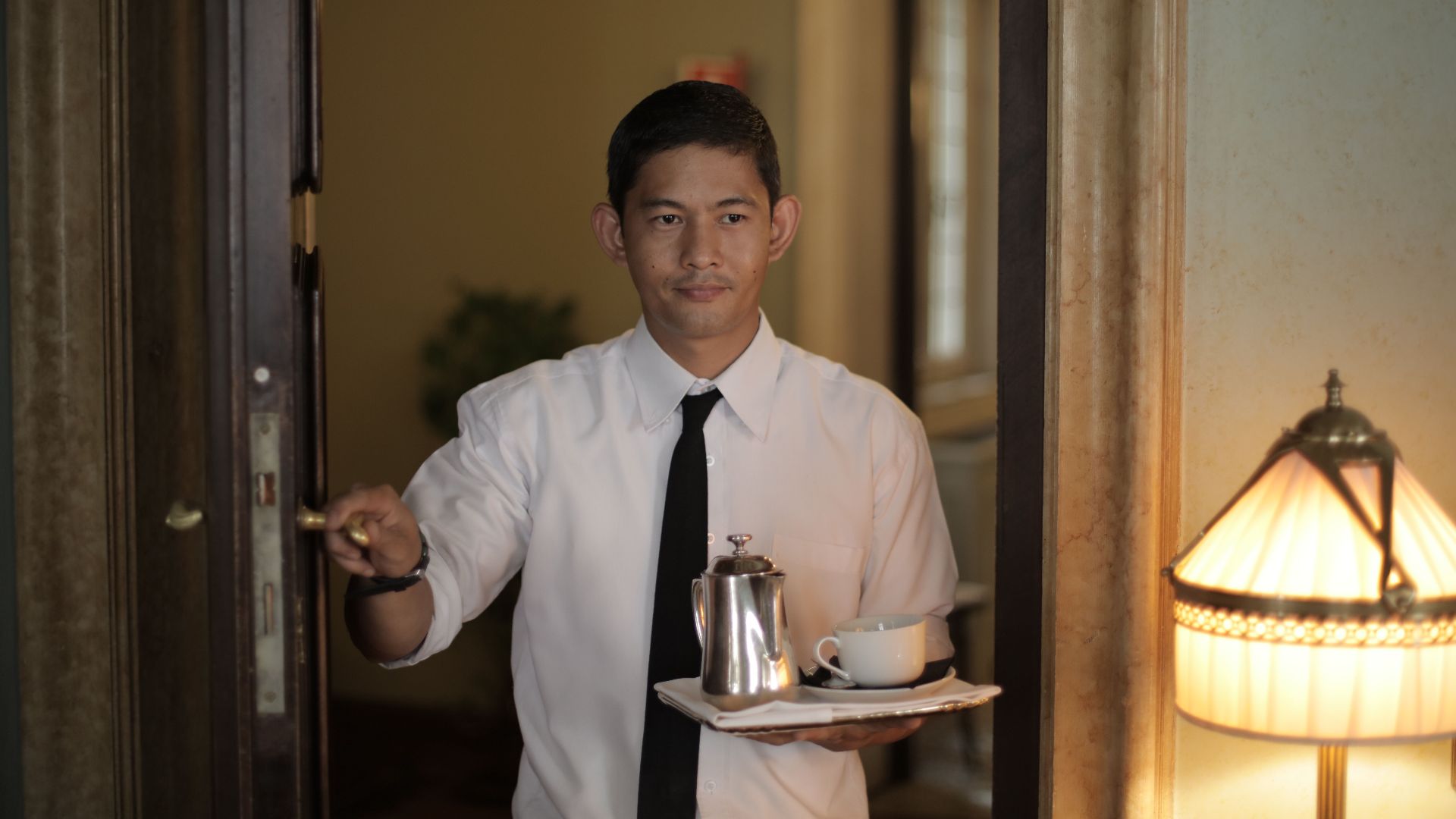 Positive ethnic male employee carrying tray with coffee while serving guest in cozy hotel room