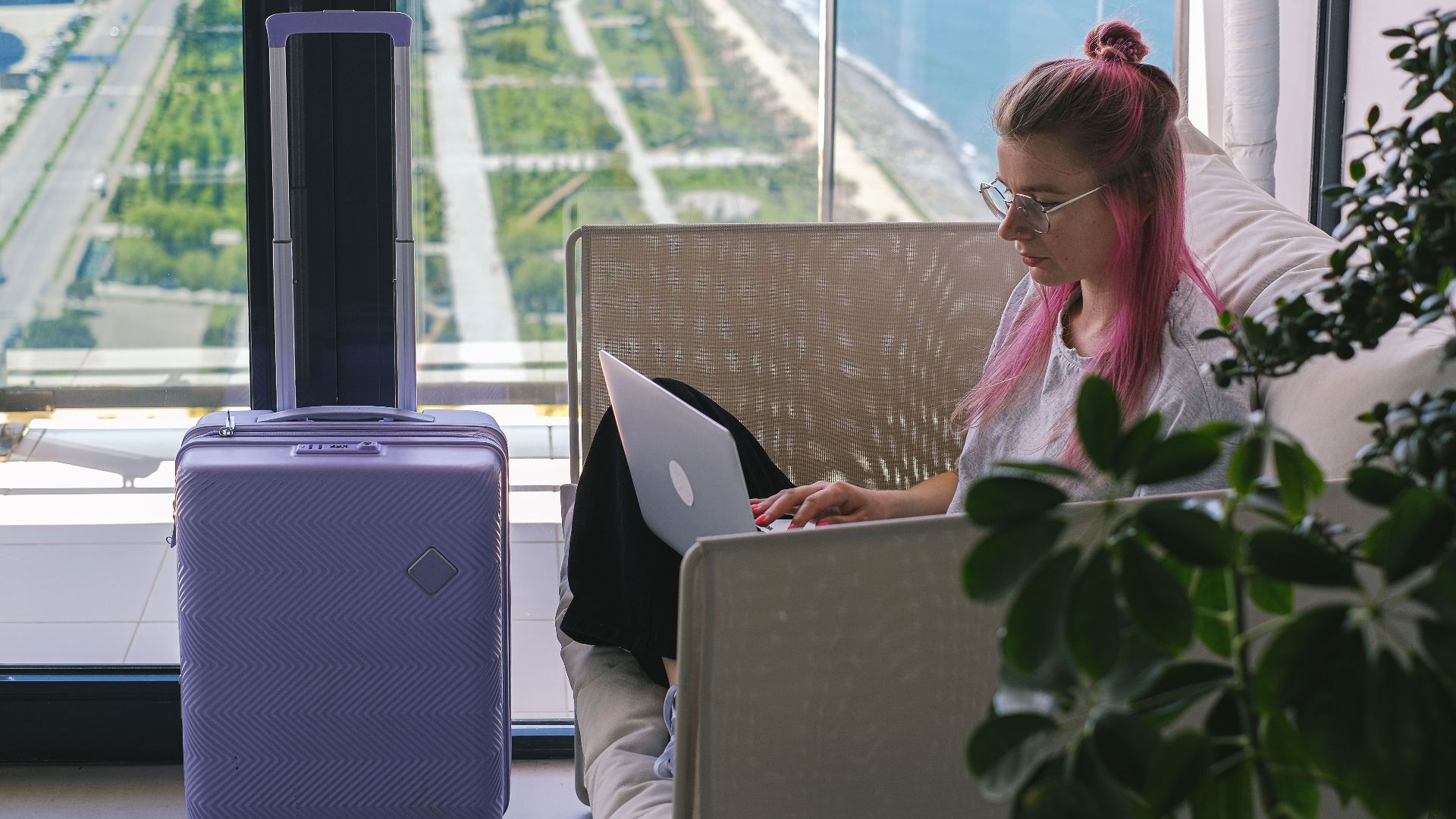 Woman with pink hair working on laptop in hotel room overlooking scenic landscape. Travel and freelance concept.