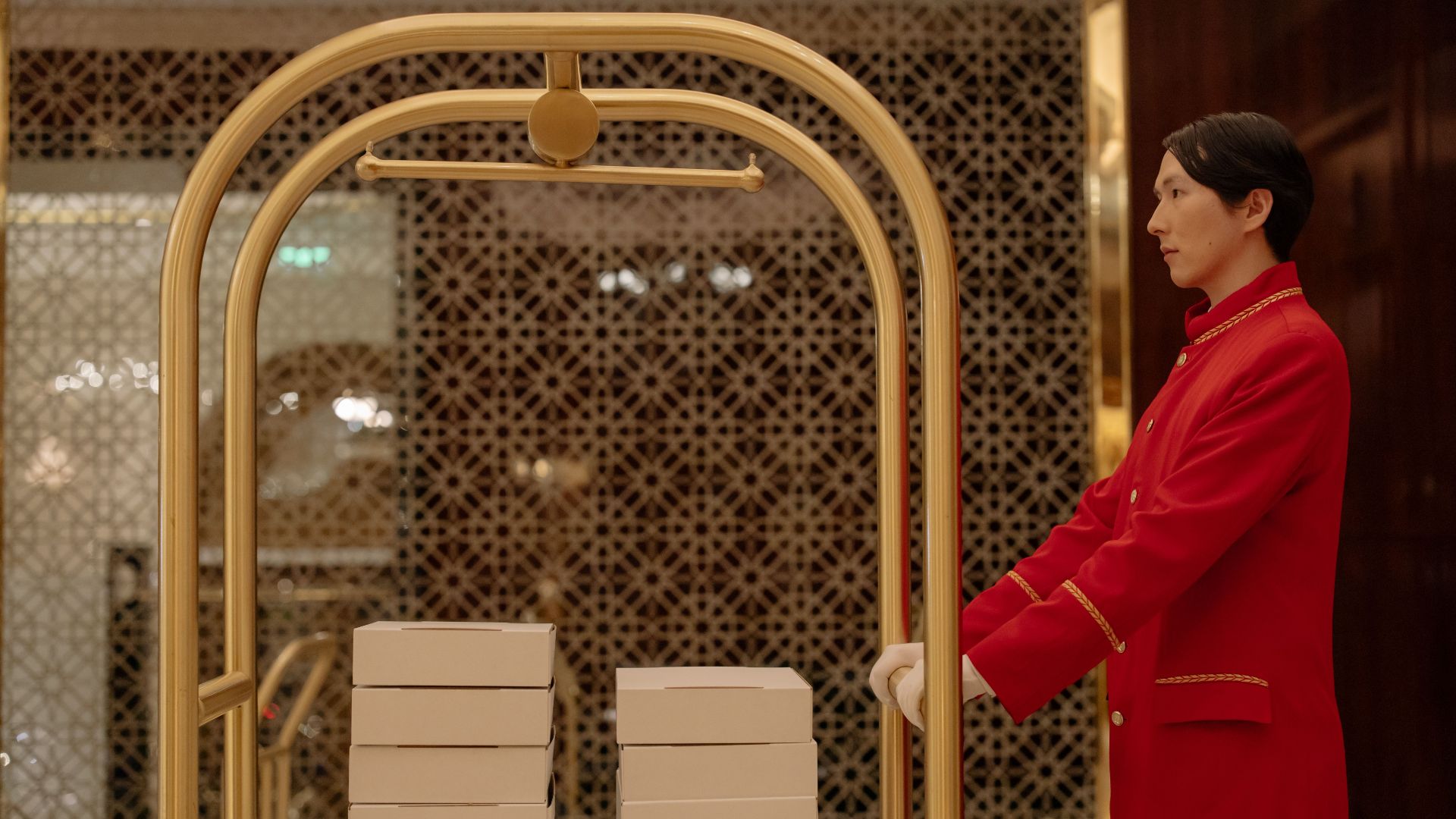 A hotel bellboy in uniform pushes a luggage cart with boxes indoors, demonstrating hospitality service.