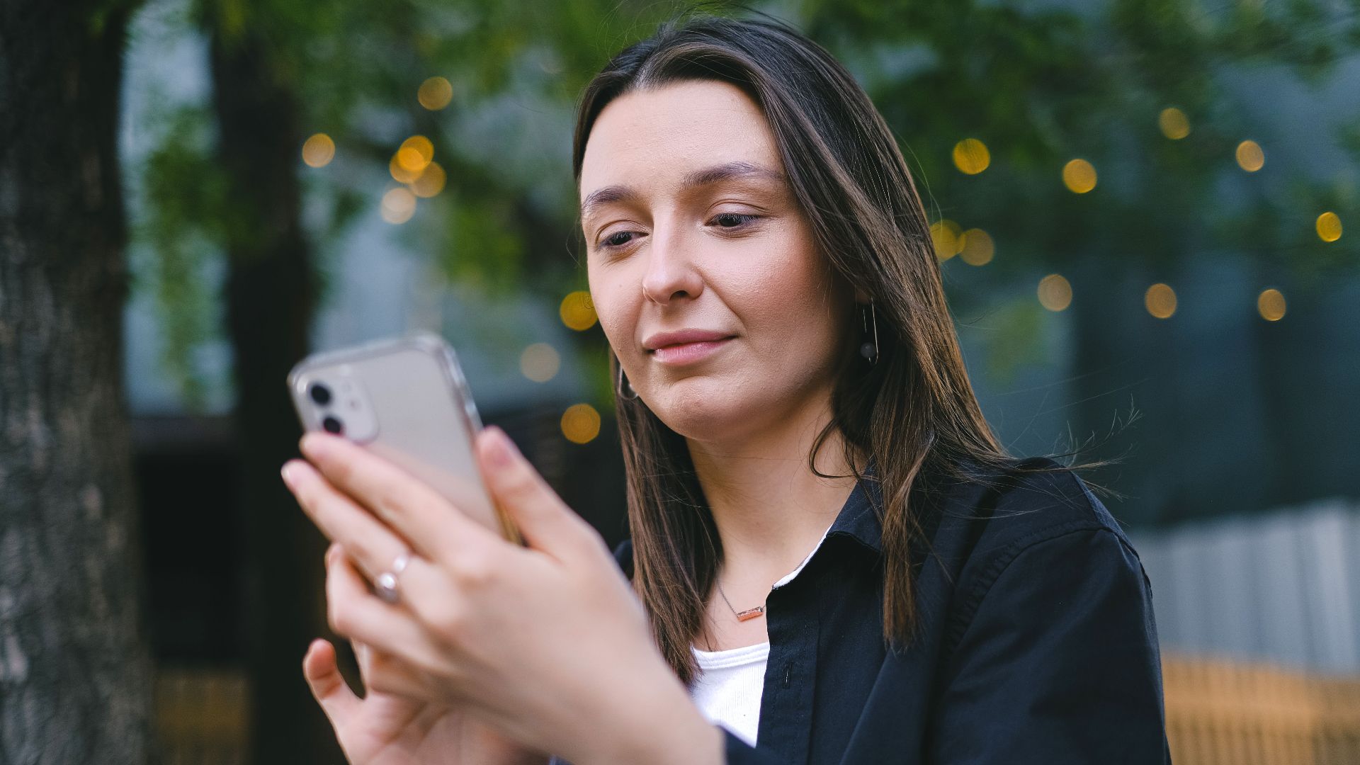 A young woman looking at her smartphone with a gentle smile in an outdoor setting, surrounded by nature.