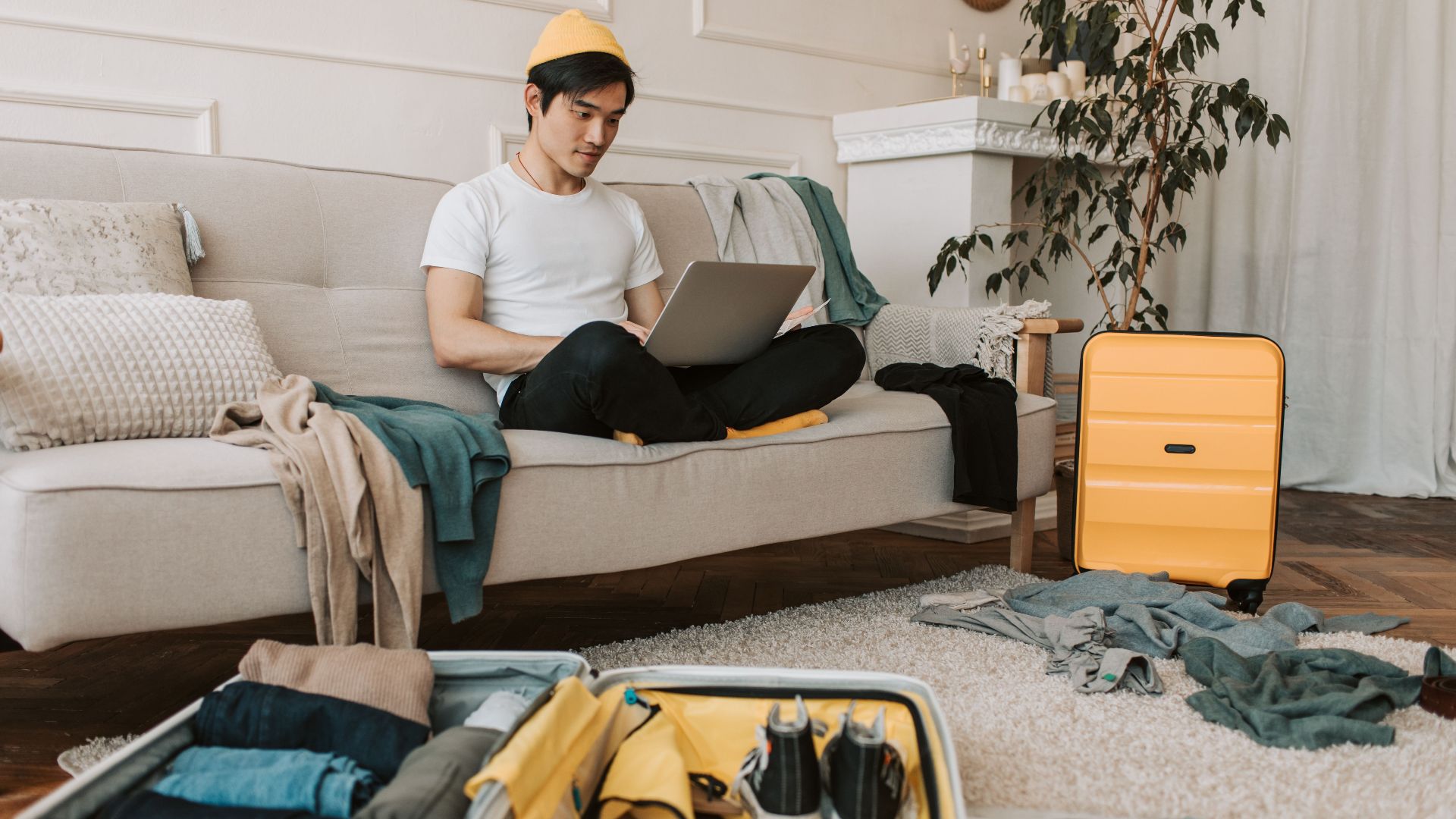 A young man in yellow accessories works on a laptop among unpacked luggage at home.