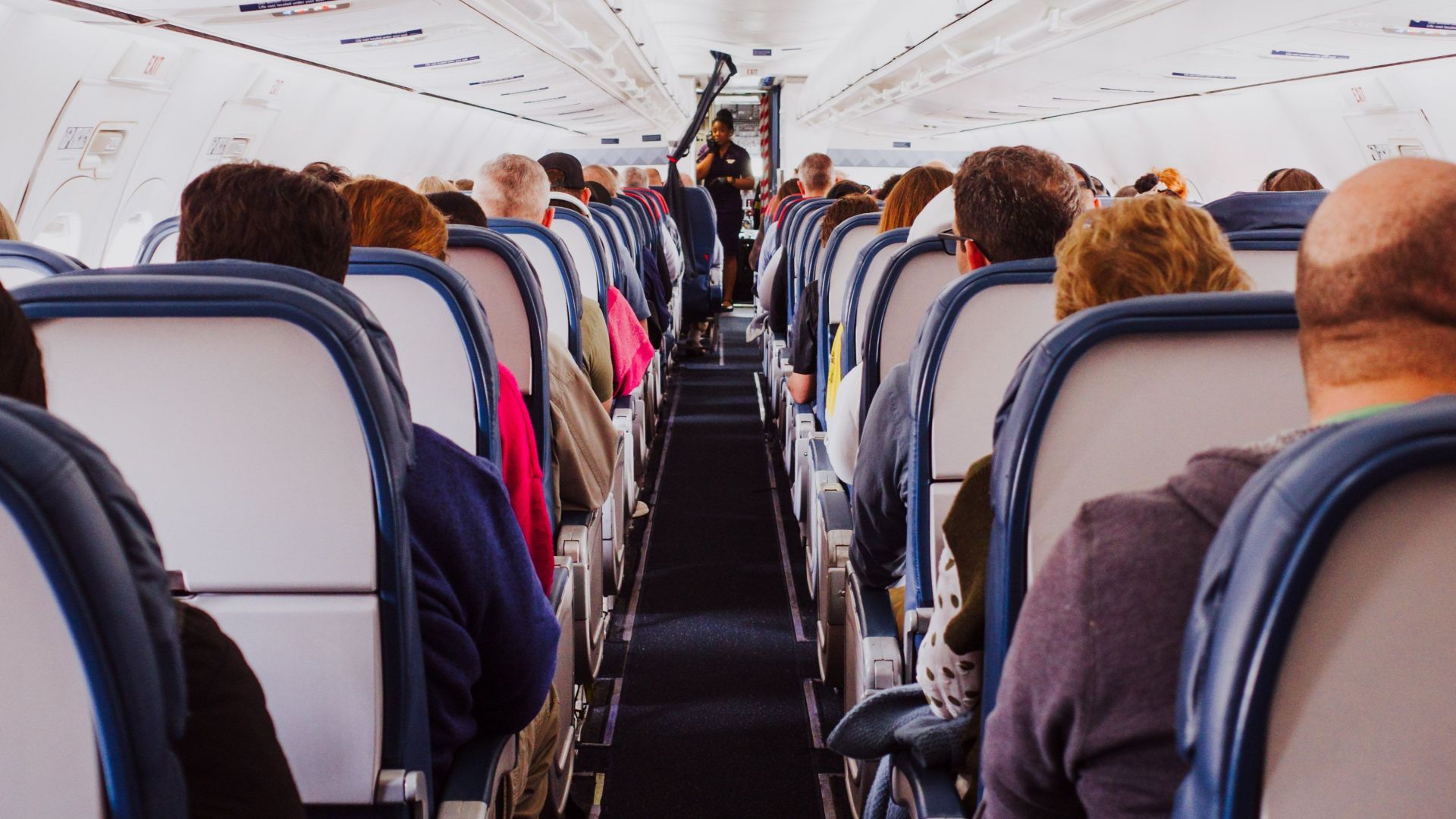 View from the back of a crowded airplane cabin showing passengers seated in rows during flight.