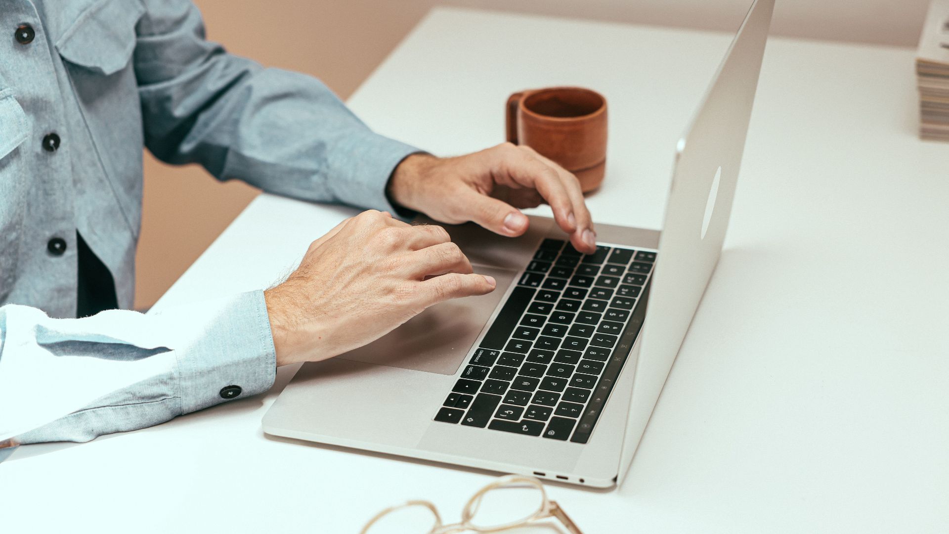 Adult working remotely on laptop with eyeglasses and cup on a white table.