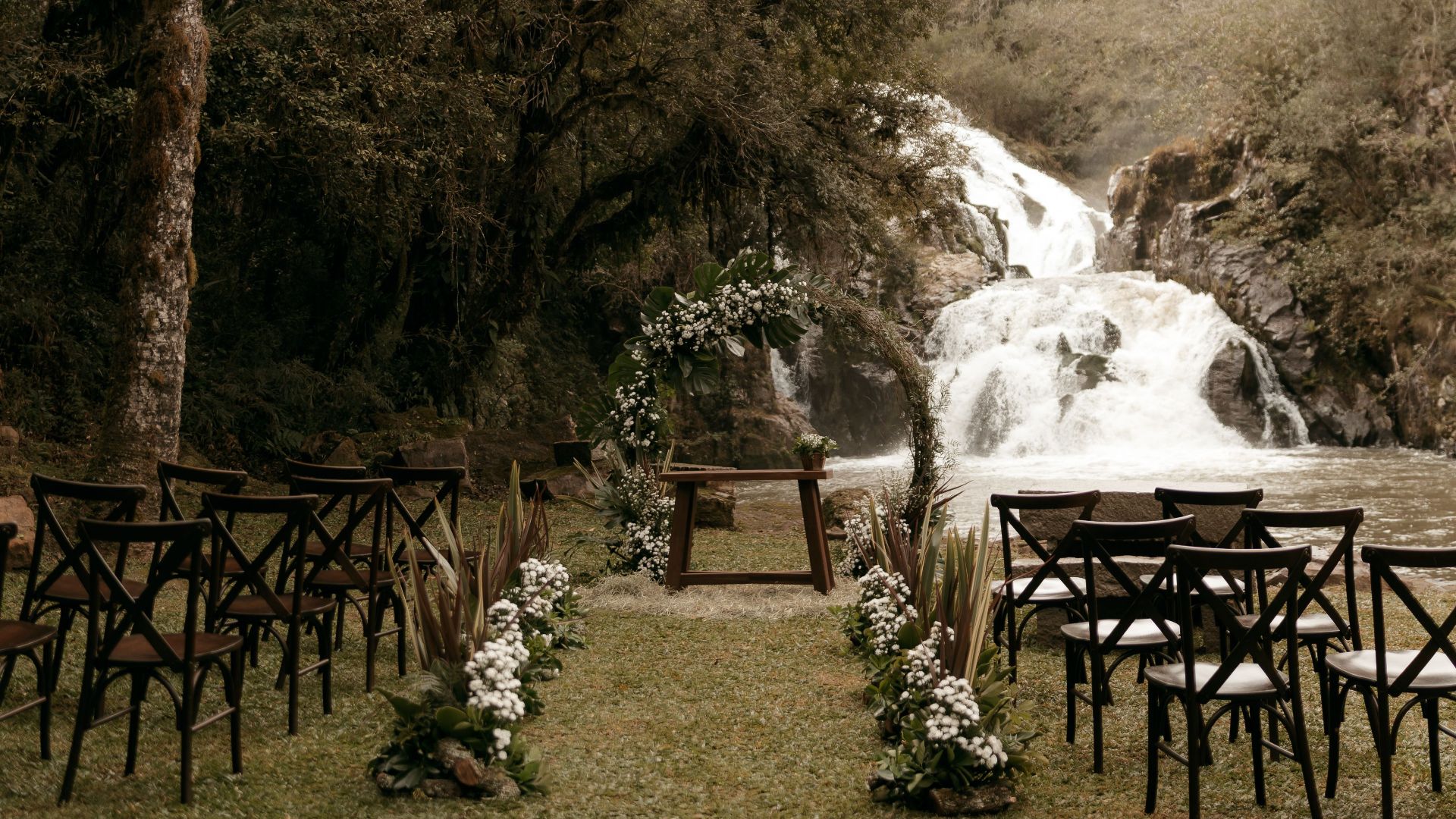 Serene outdoor wedding setup by a picturesque waterfall with rustic wooden chairs and floral decor.