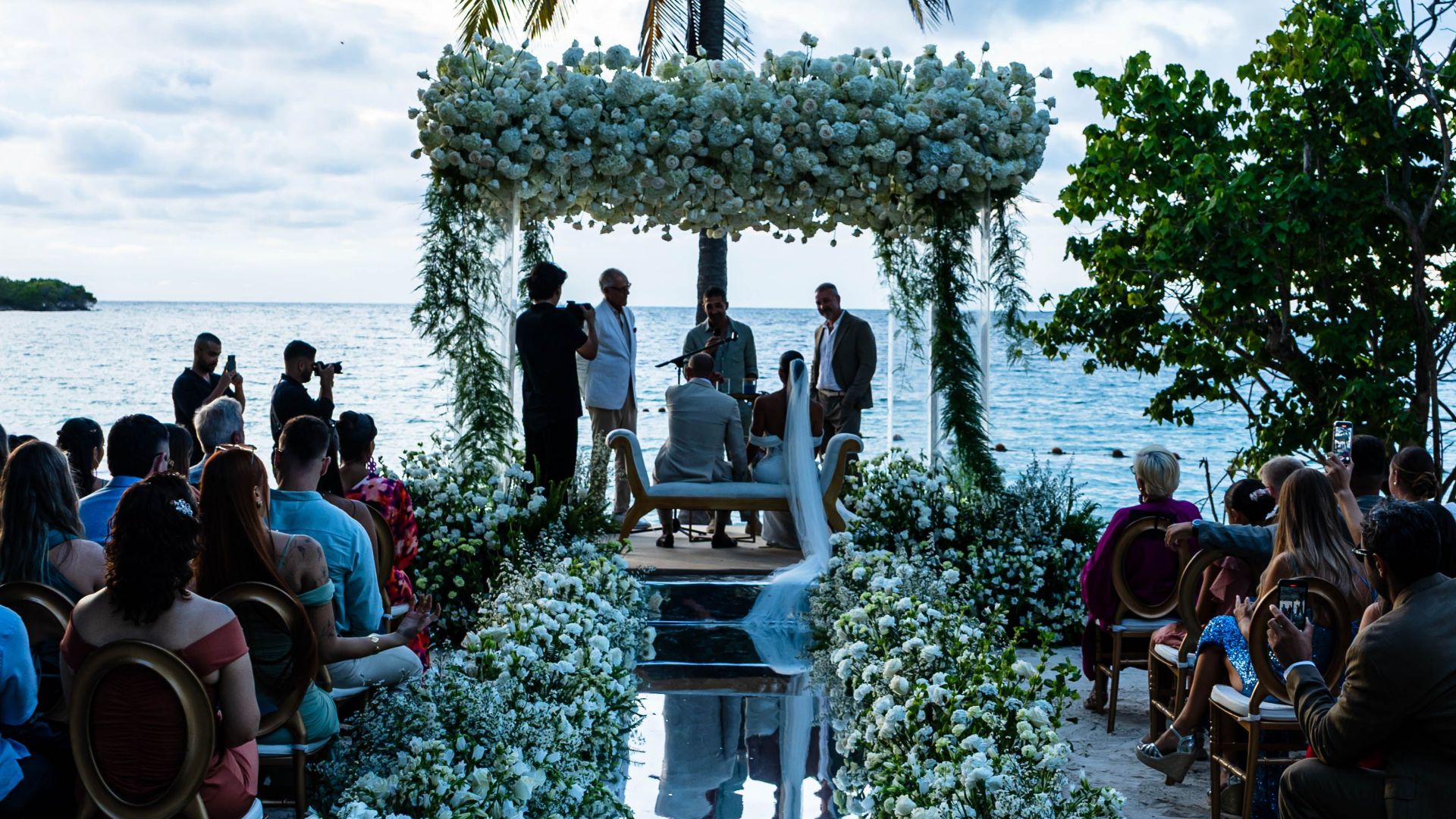Romantic wedding ceremony on a tropical beach at sunset with palm trees and floral decorations.