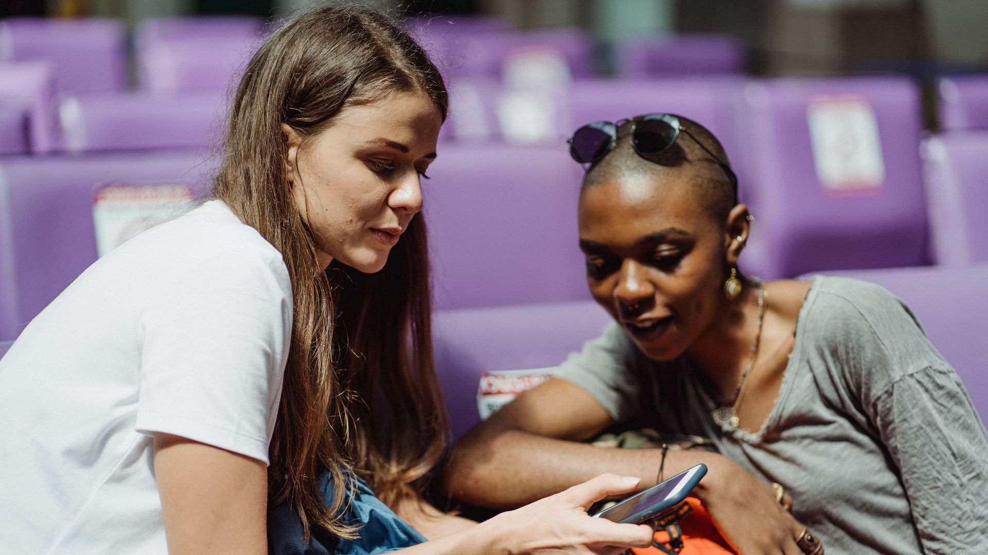 Two adult women sitting together and sharing a smartphone in a public waiting area with purple seats.