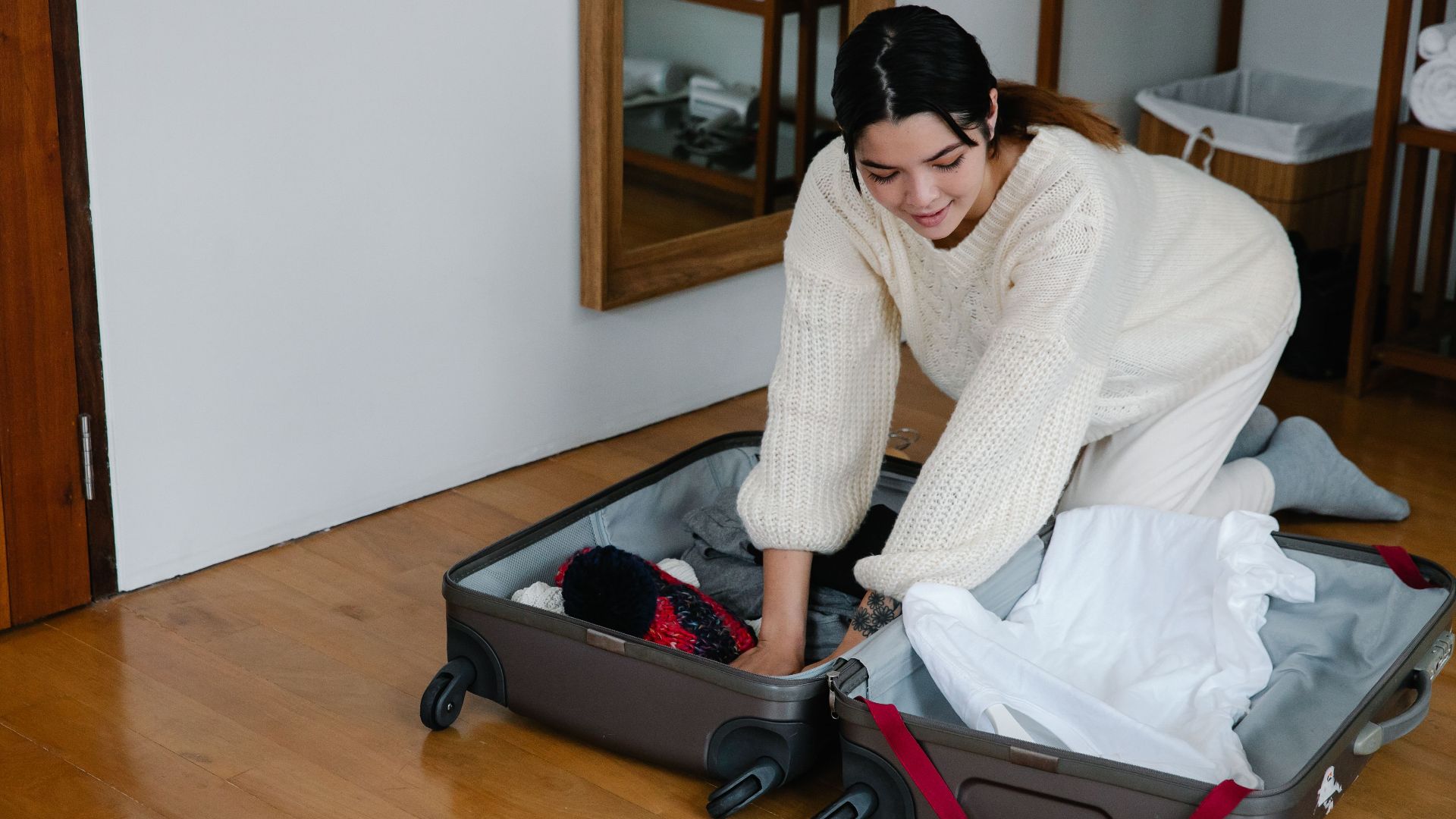 A woman kneels on the floor packing clothes into a suitcase in a cozy bedroom setting.