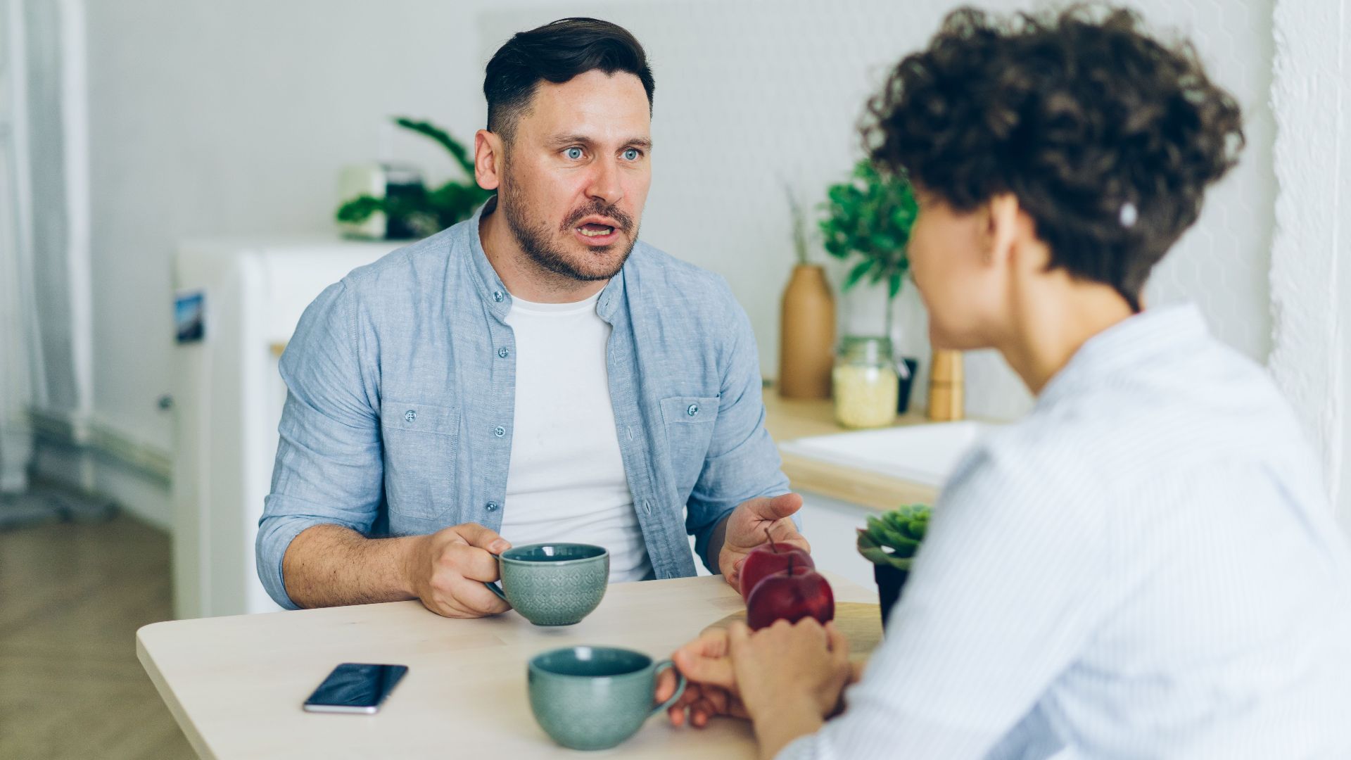 Couple having a serious conversation at the kitchen table with coffee cups.