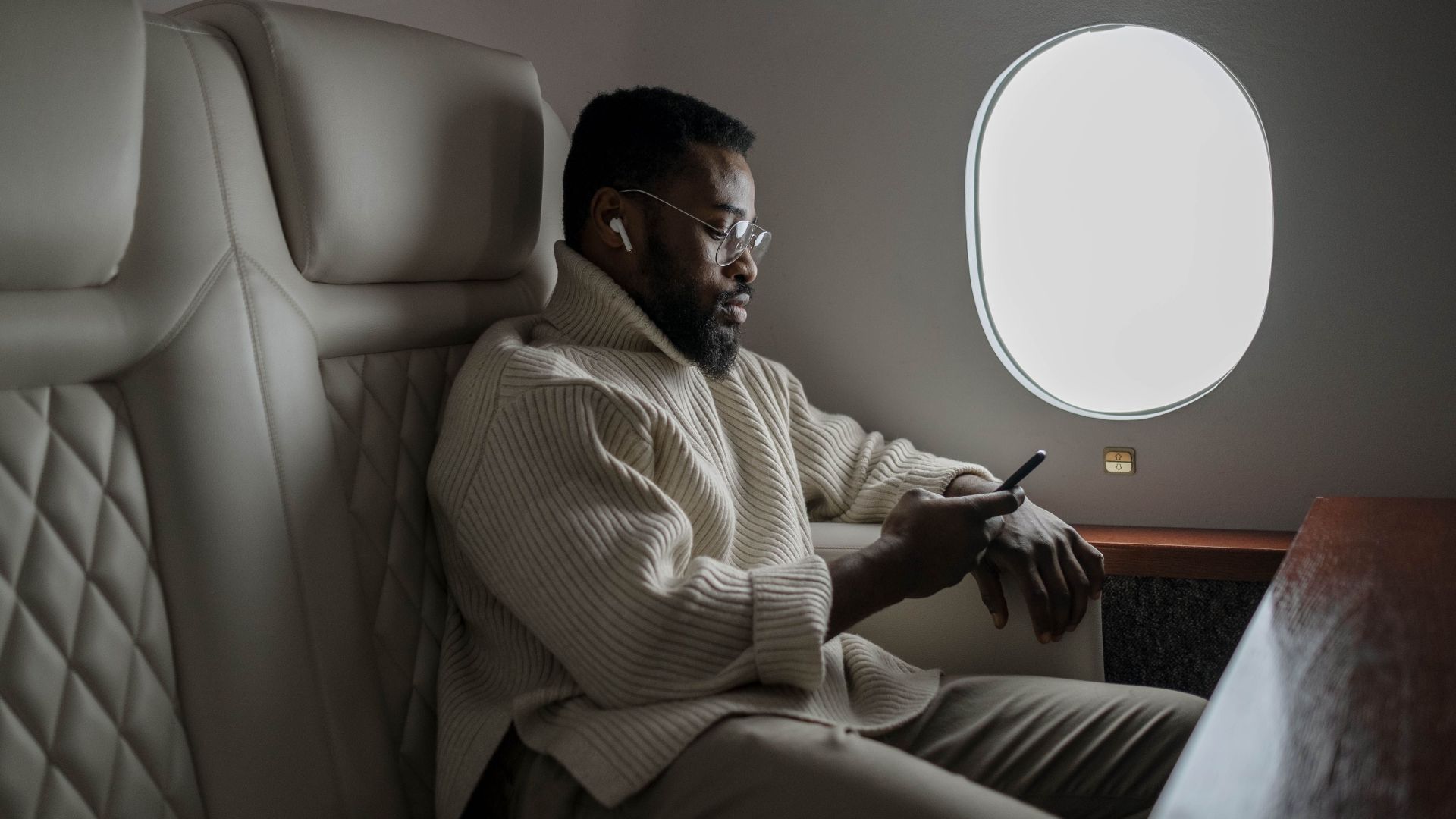 Bearded man in cozy sweater using smartphone on airplane, enjoying in-flight entertainment.