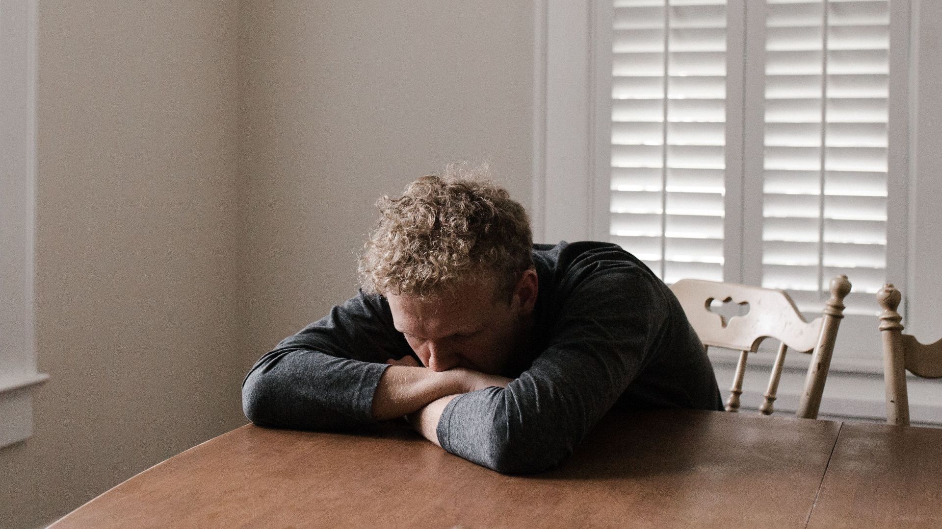 An adult man with emotions of sadness and anxiety sitting alone at a wooden table near windows.