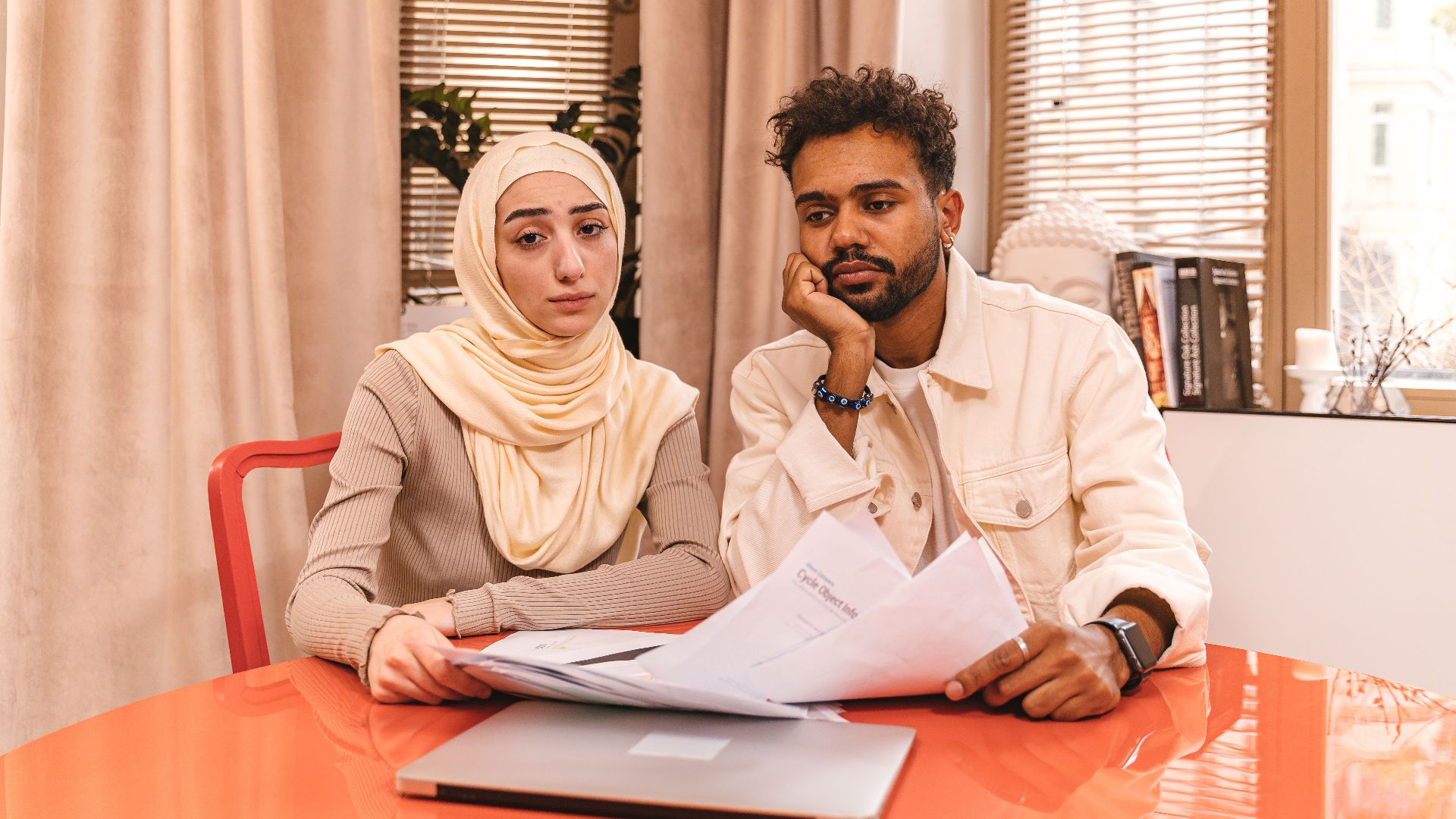 A couple in casual attire sits indoors at a table reviewing financial documents with a laptop.