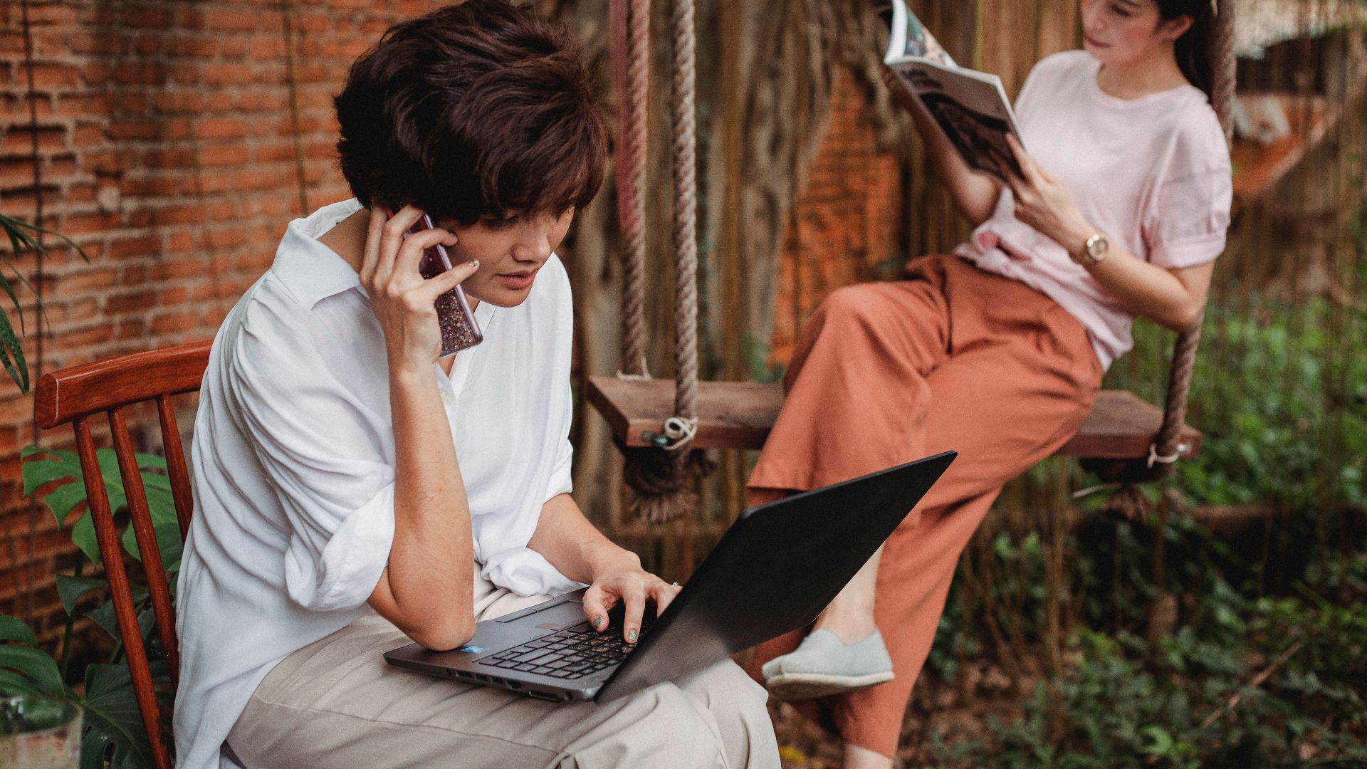 Pensive Asian female browsing laptop and talking on smartphone near resting Asian female reading magazine in garden near brick building in daytime