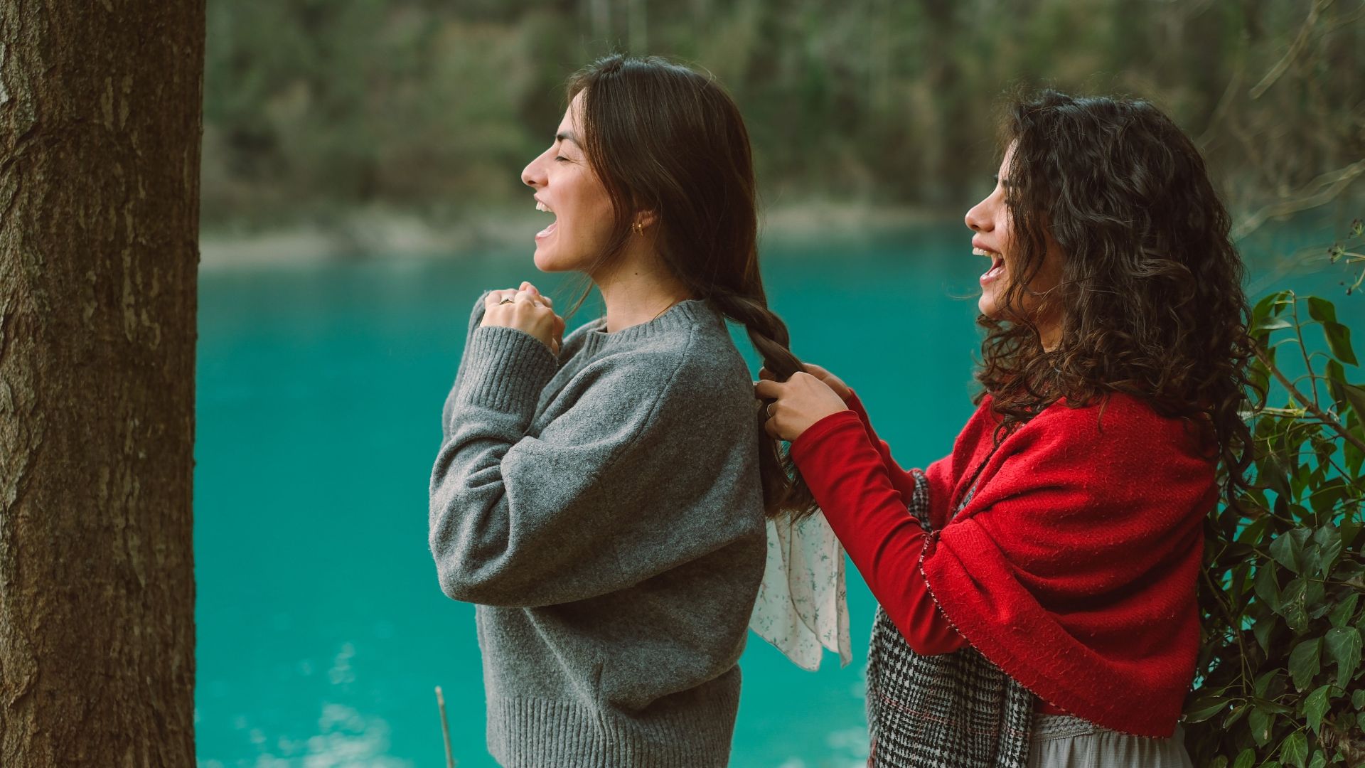 Two women enjoying a playful moment by a turquoise lake, styling hair amidst nature.