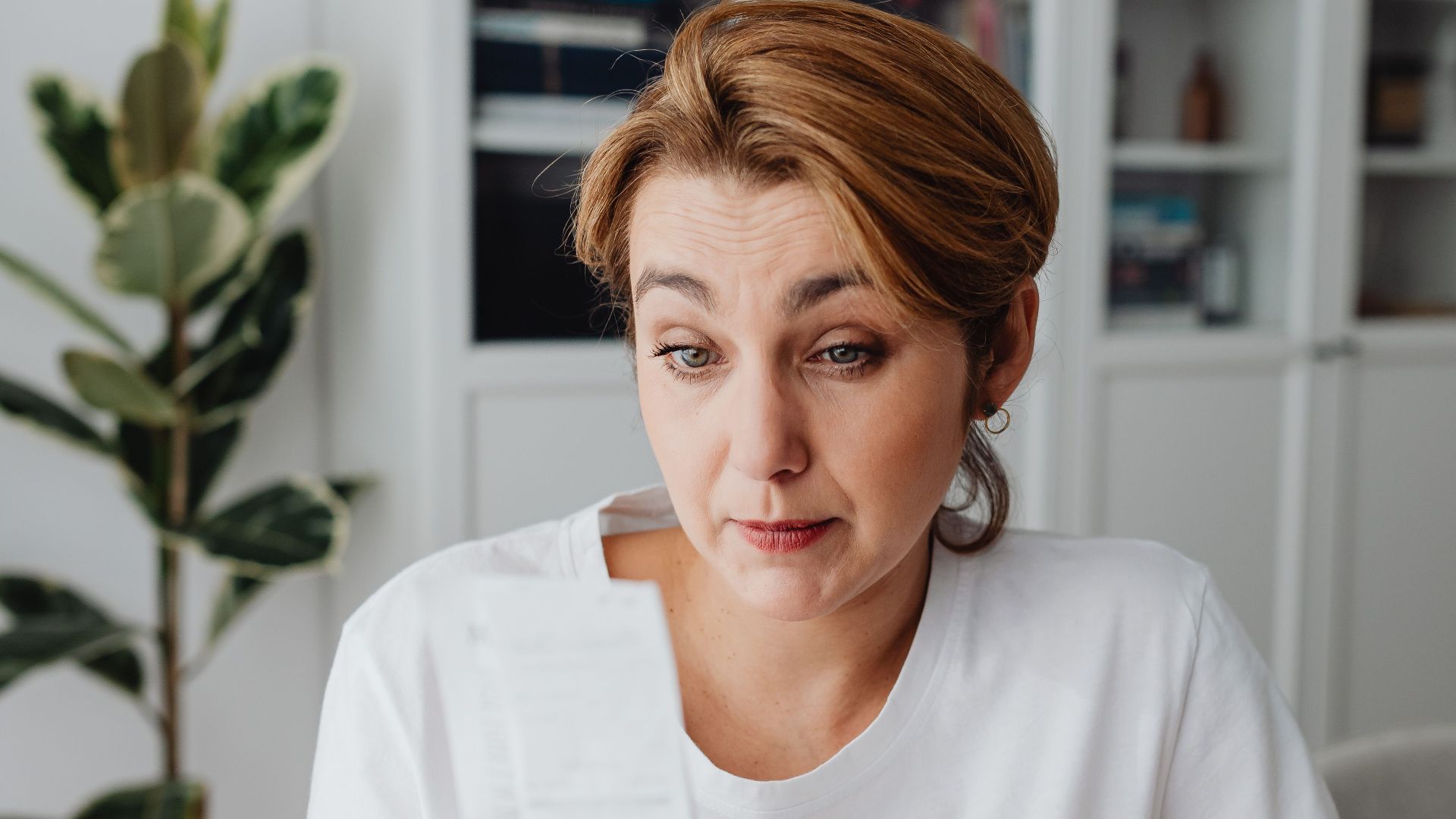 An adult woman with a surprised expression looks at a receipt at her desk.