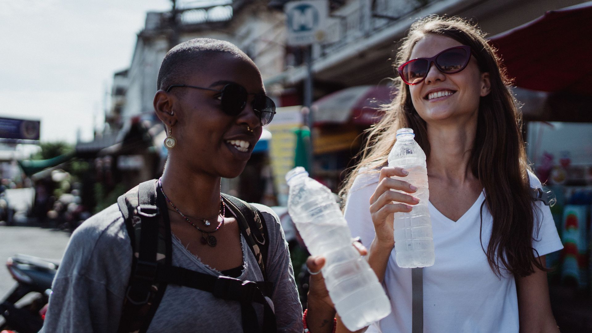 Two young women enjoying a city adventure, holding water bottles, and smiling under the summer sun.