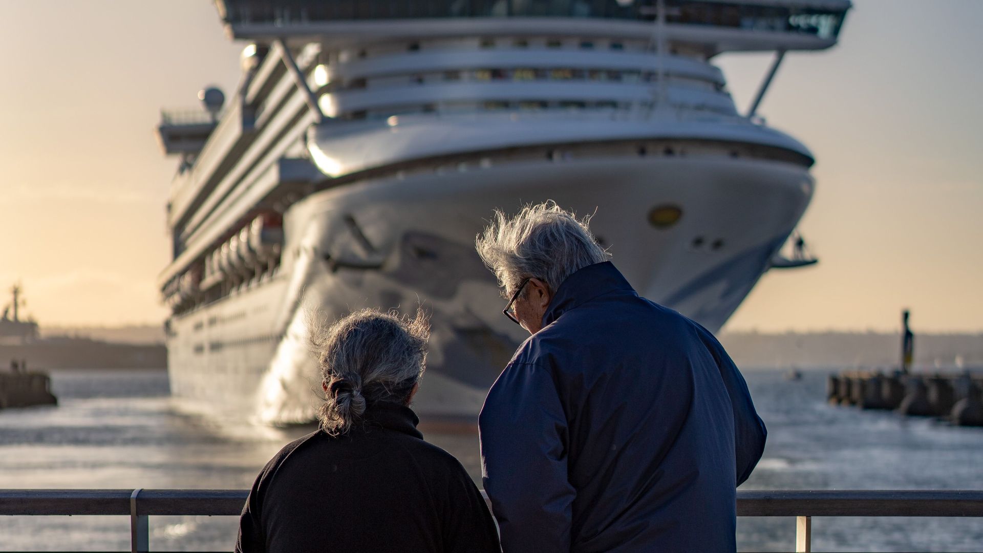 Senior couple standing at railing, admiring large cruise ship approaching dock at sunset.