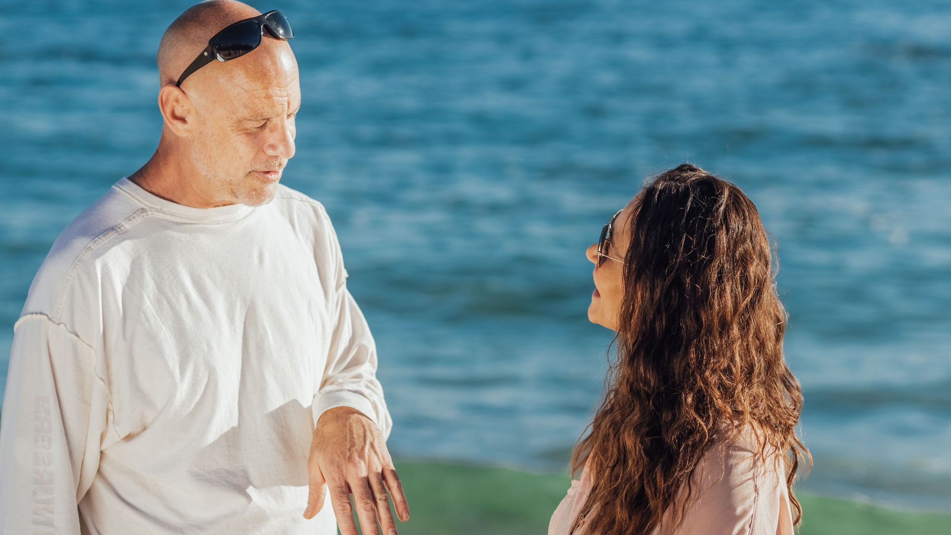 Two adults enjoy a casual conversation on a sunny beach with the ocean as a backdrop.