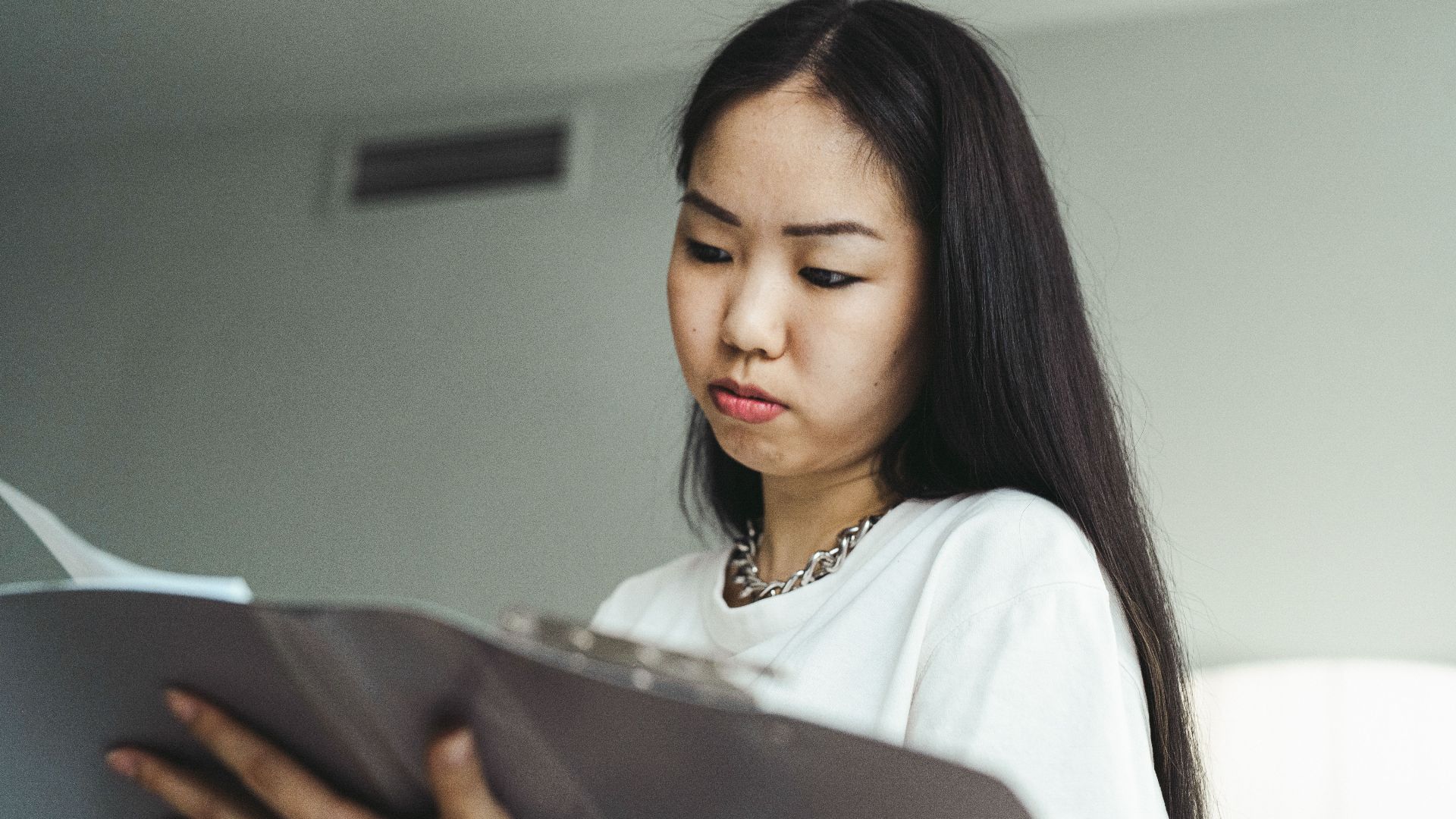 Asian woman in casual attire examining paperwork with concentration indoors.