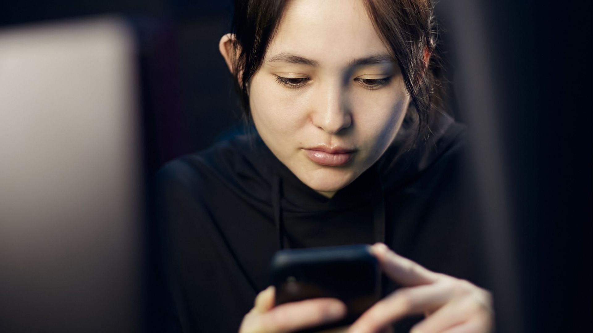 A woman intently looking at her smartphone while seated indoors, dim lighting.