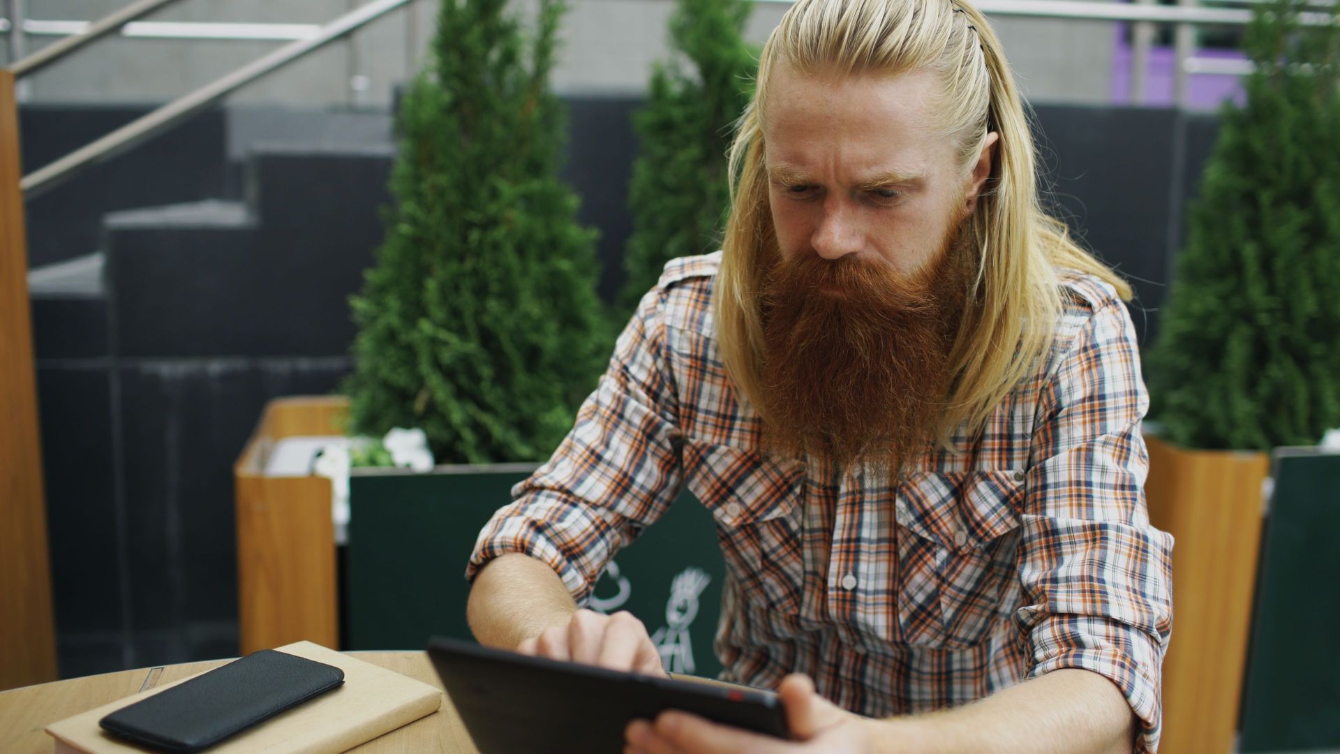Focused man with long hair and beard using a tablet at an outdoor cafe setting.