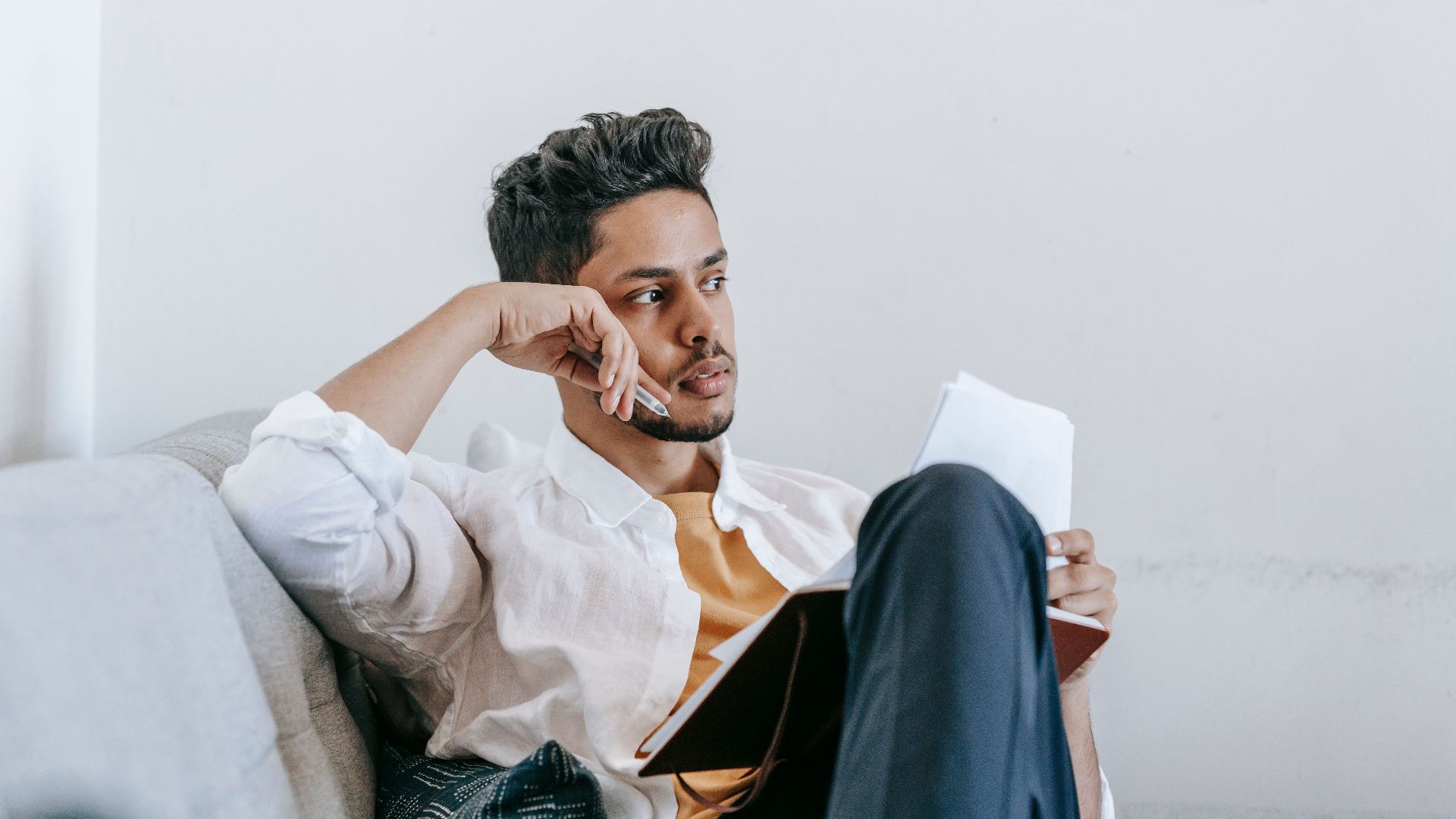 Contemplative young ethnic male in casual wear reading notes in planner and looking away in thoughts while resting on comfortable couch
