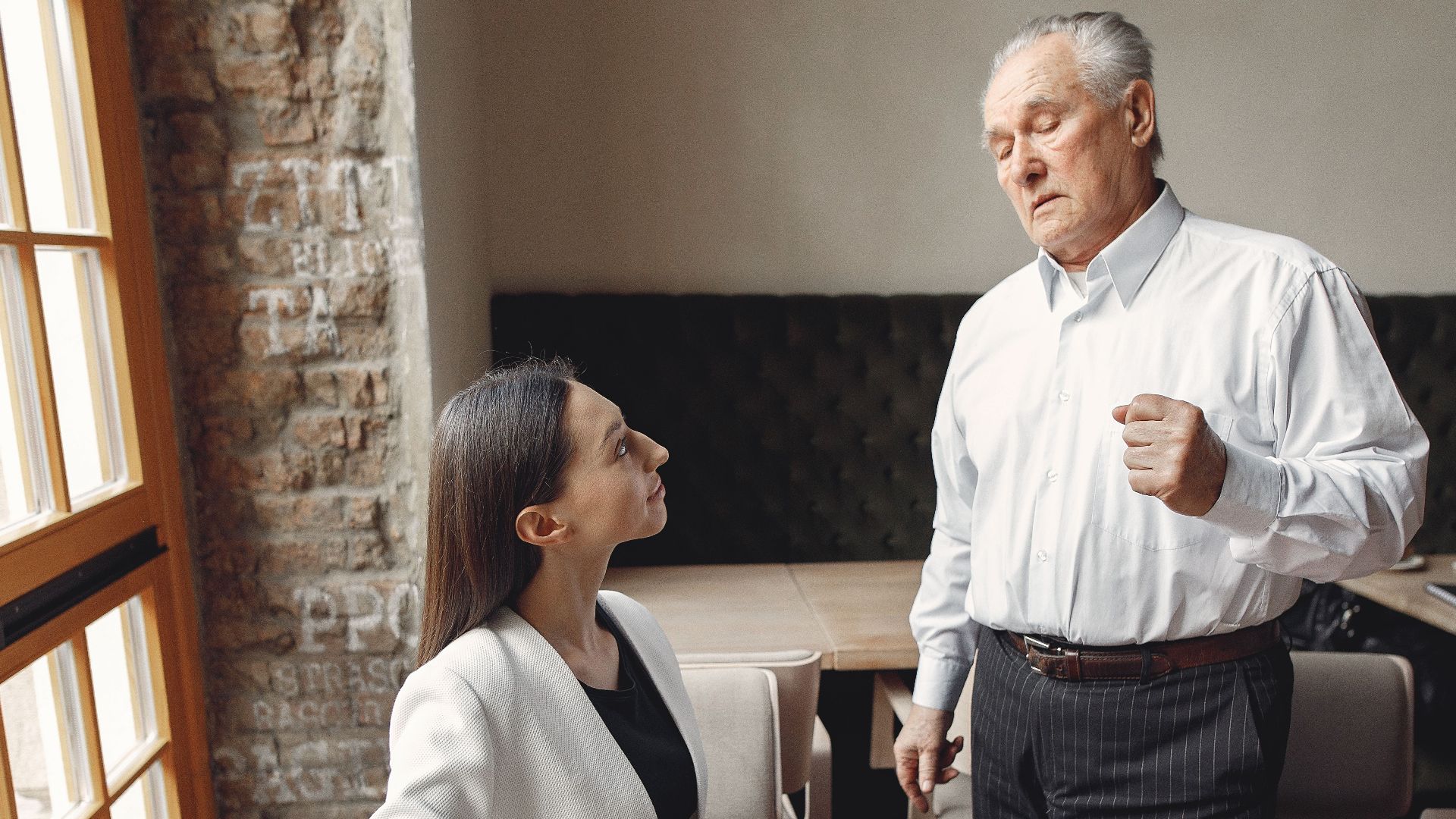 Aged businessman in formal wear talking with well dressed female employee with laptop and cup of coffee about business issue in modern office