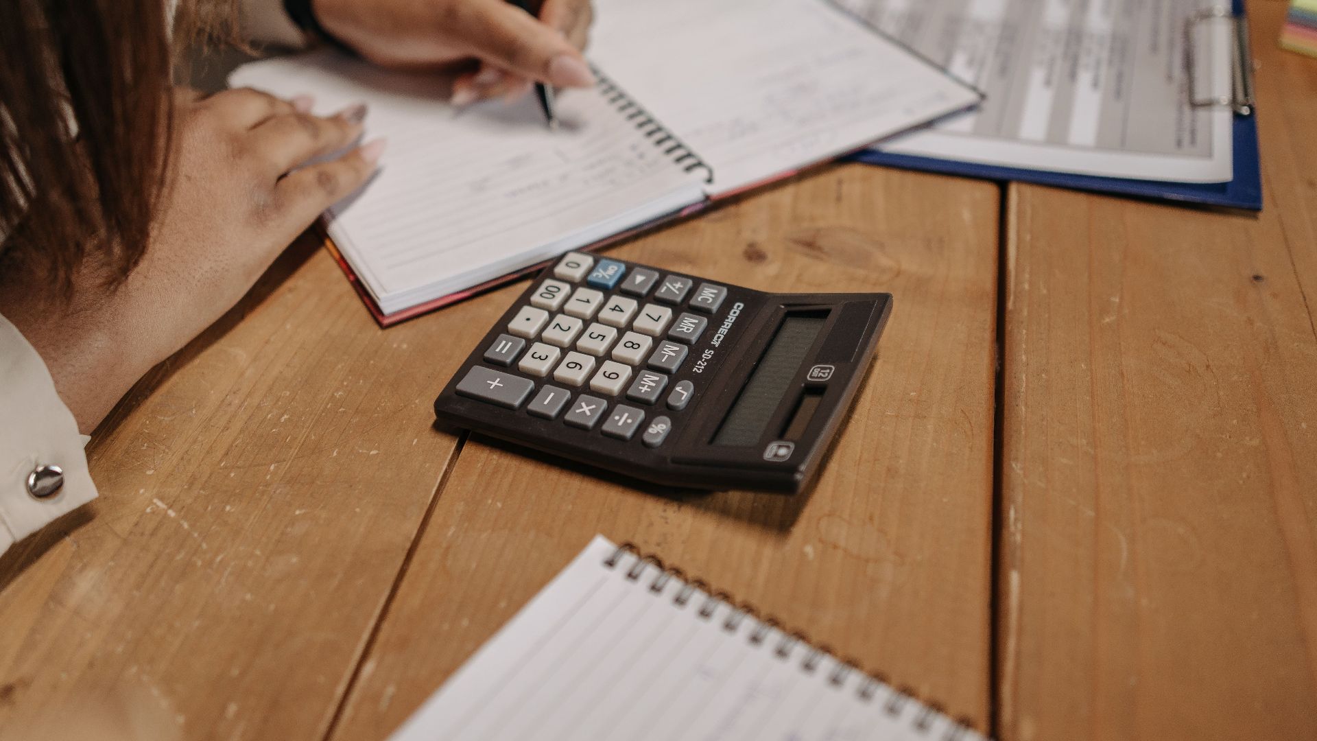 Woman writing notes with a calculator on a wooden desk, focusing on work and accounting.
