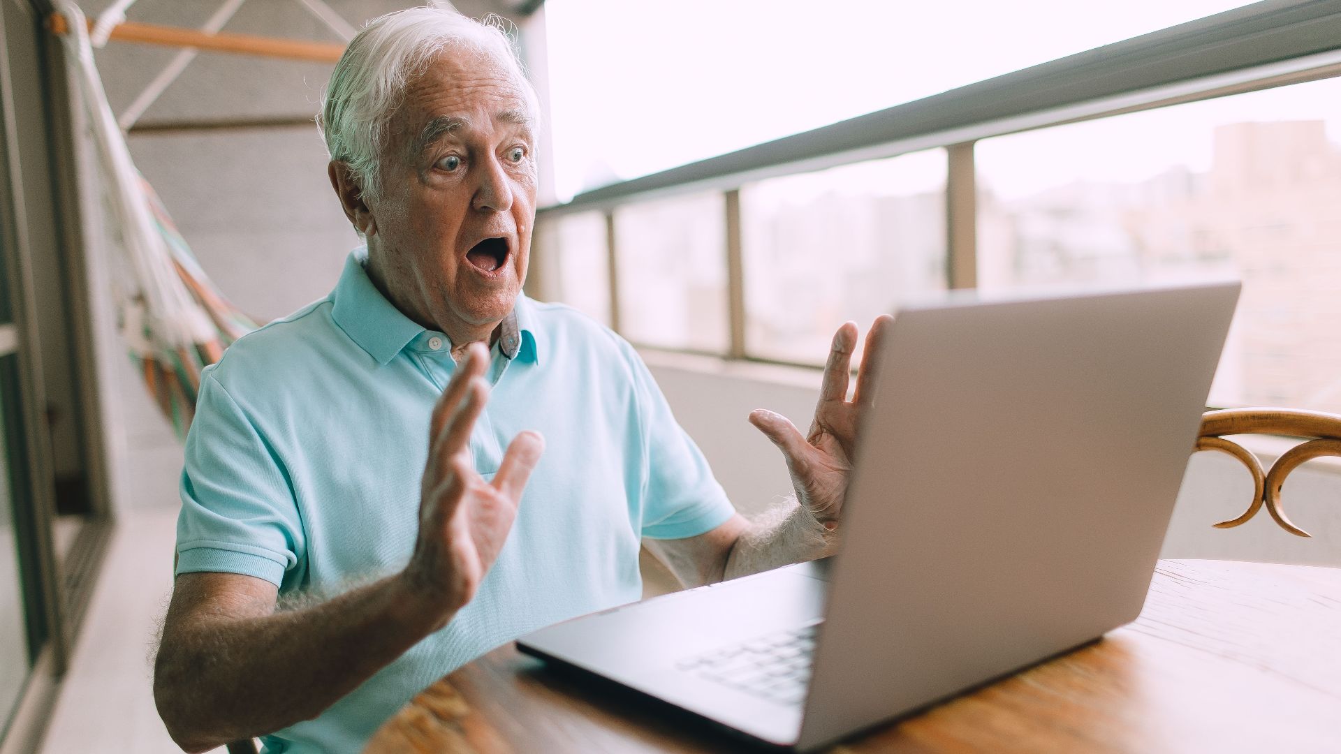 Elderly man looking shocked while using a laptop on a balcony, depicting excitement or surprise.