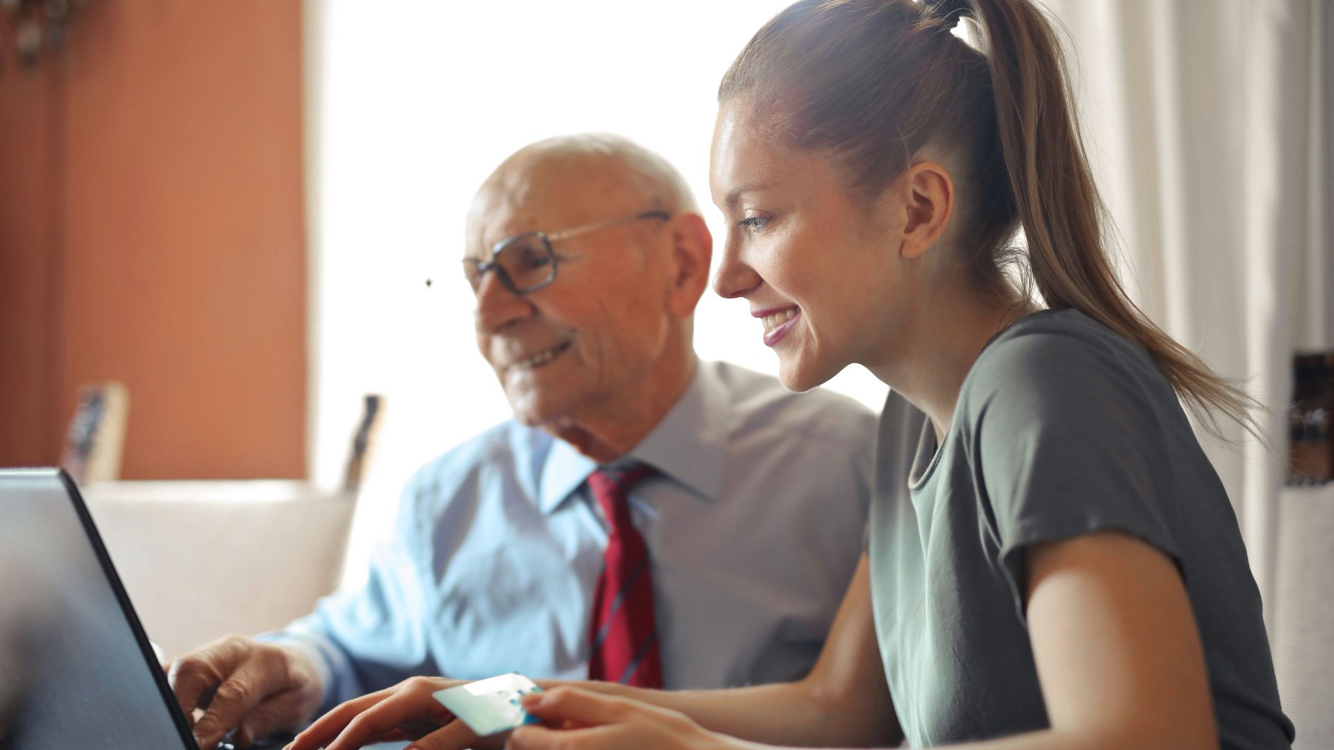Young woman in casual clothes helping senior man in formal shirt with paying credit card in Internet using laptop while sitting at table