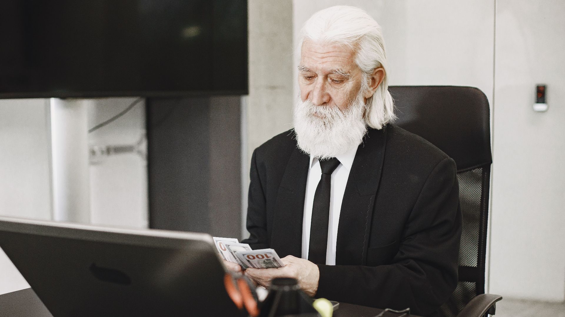 White-haired man in suit counting money at a modern office desk with laptop.