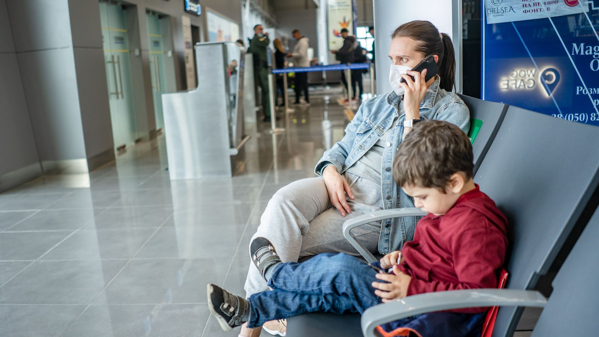 A mother and child sitting at an airport terminal. The mother is on the phone while the child is using a device.