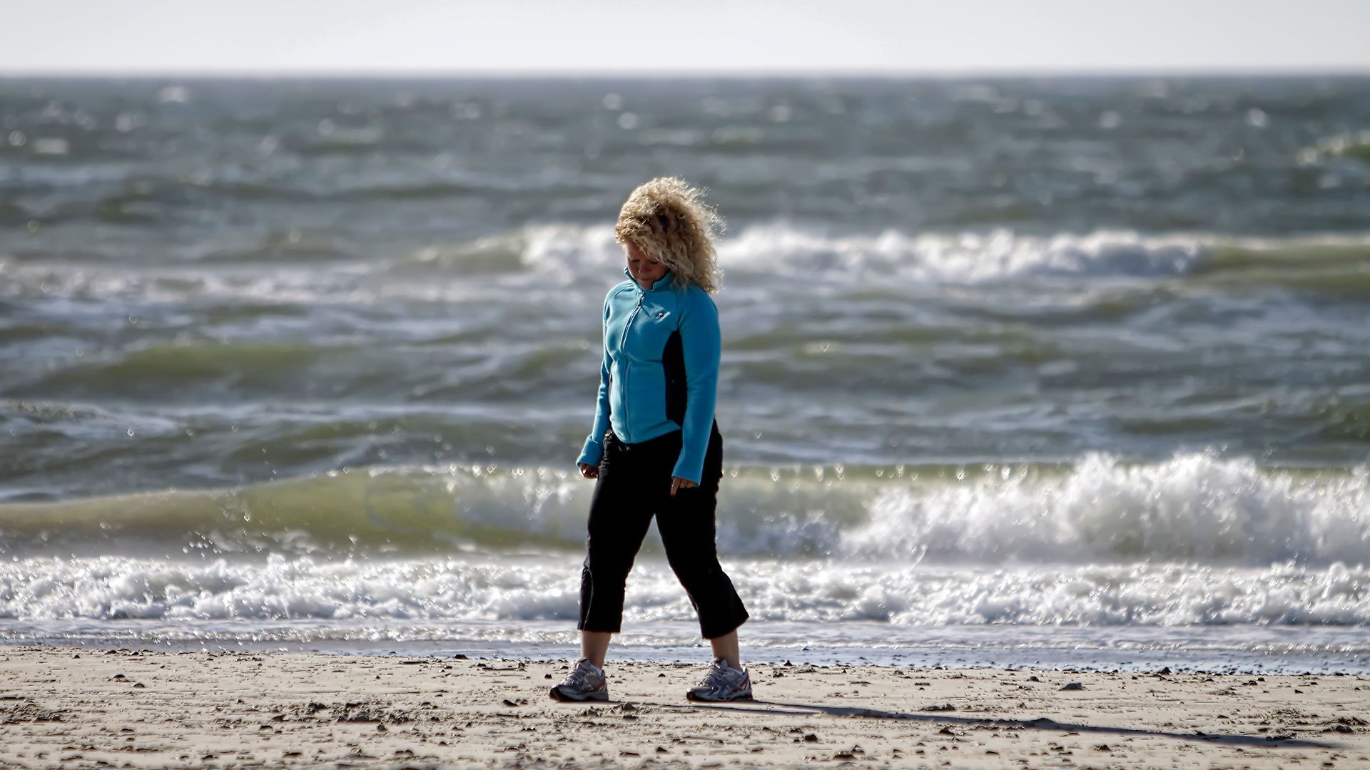 A woman in activewear walks along a windy beach in Denmark, with ocean waves crashing behind her.