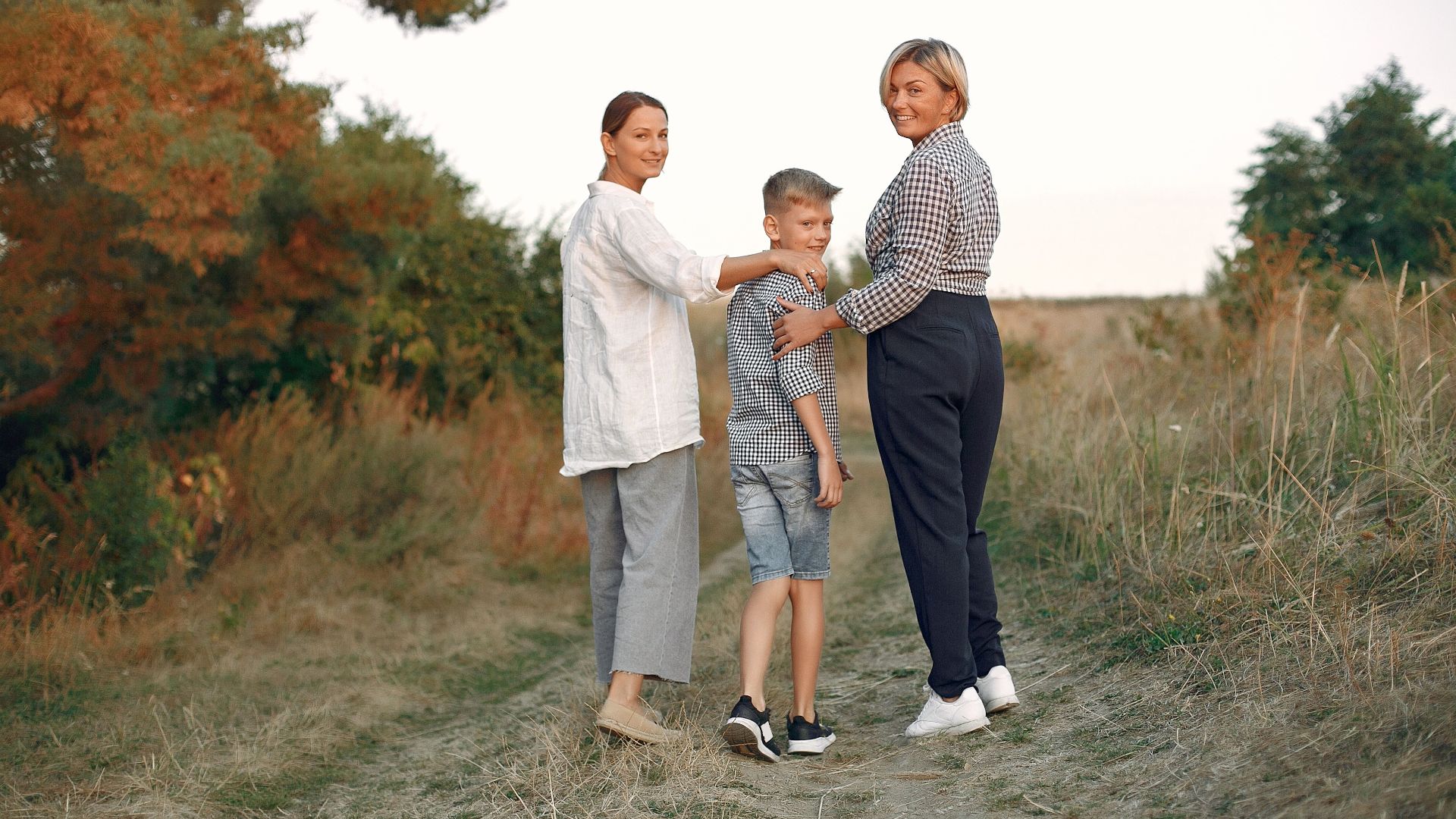 Back view of delighted adult mother and preteen son strolling together with middle age grandmother on path in summer field during weekend in countryside while looking over shoulder