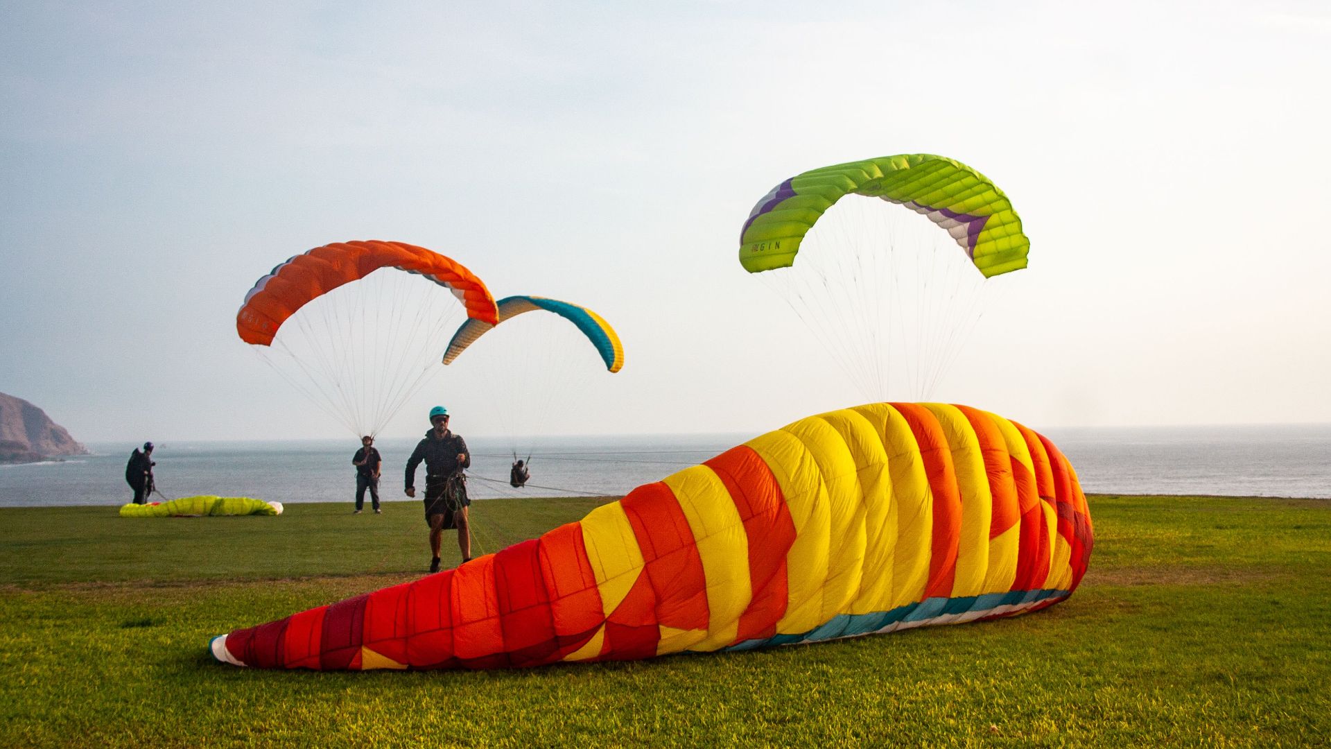 Colorful paragliders get ready to soar on a grassy coastal hill with a scenic sea view.