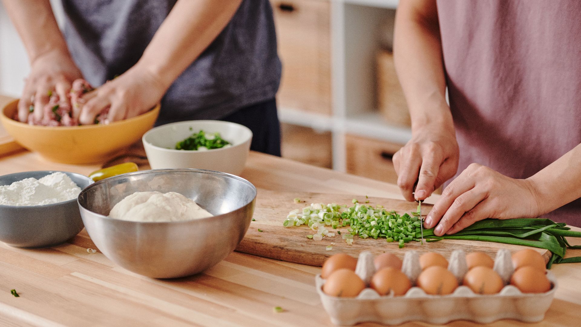 Two people preparing food with fresh ingredients on a wooden table in a cozy kitchen environment.