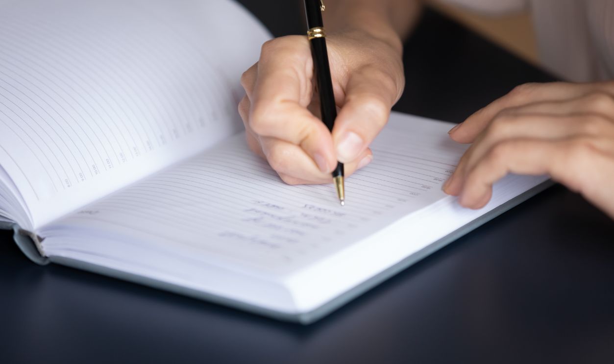 Housewife holding pen creates household to-do list using personal organizer seated at table, close up view. Businesswoman writing on diary, keeps daily record of events, appointment. Wish list concept