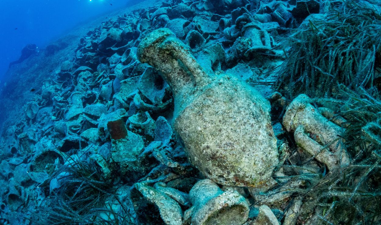 Underwater scene of an ancient shipwreck site scattered with amphorae and artifacts, covered in marine growth. The image captures the mystery of historical relics on the ocean floor