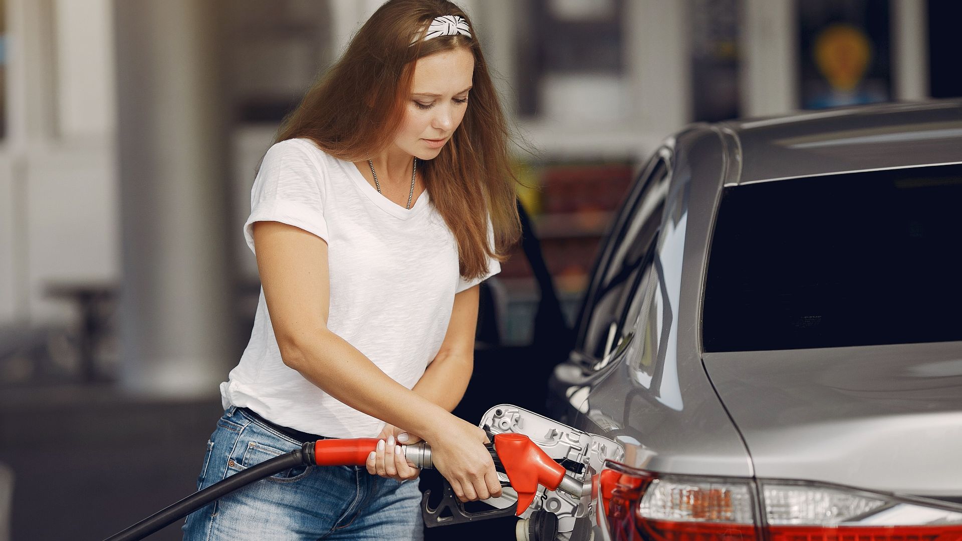 Attentive female driver in casual outfit and headband filling up modern automobile with automotive fuel gun on petrol station while looking down