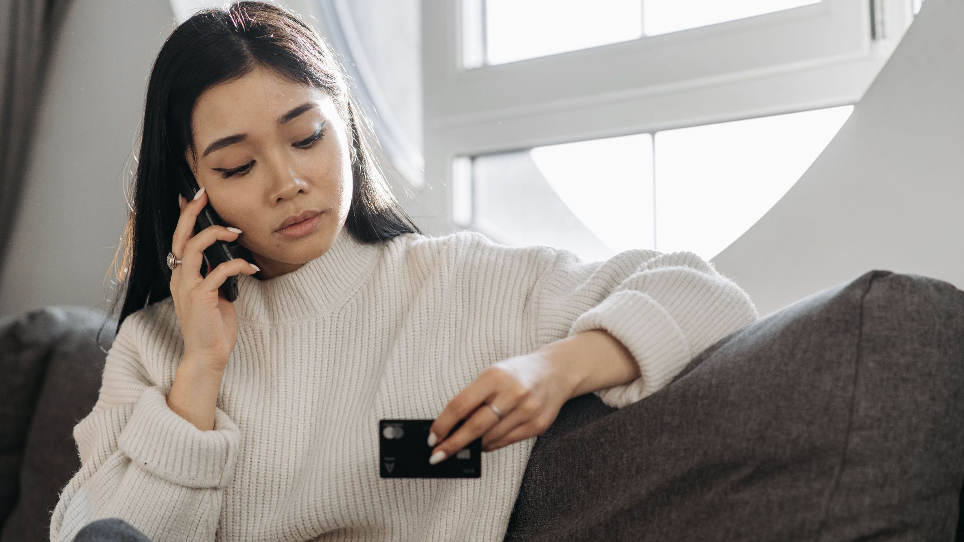 A woman sits on a sofa, multitasking with a phone and credit card, focusing on online shopping.