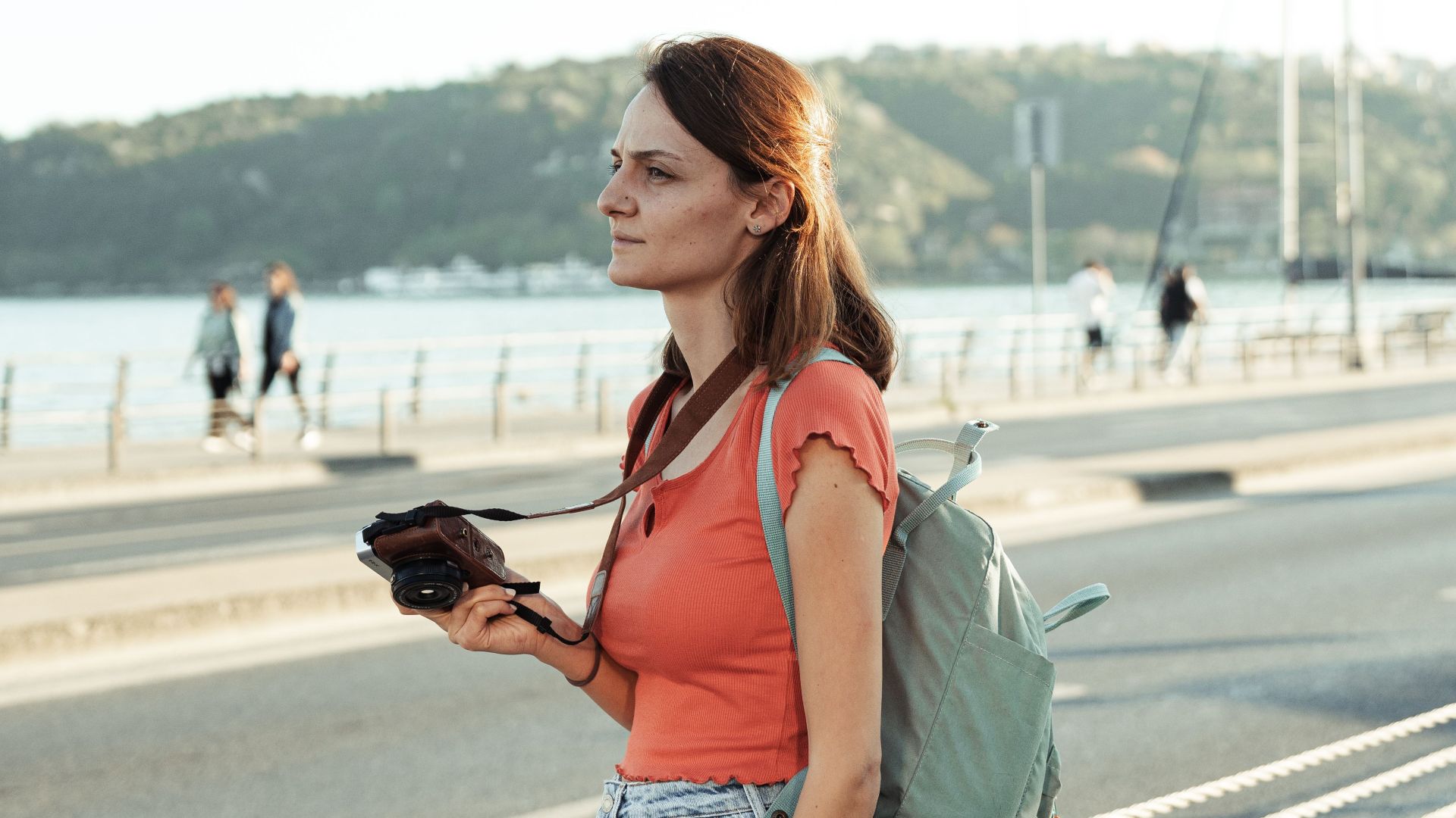 Woman standing by the waterfront with a camera and backpack on a sunny day.