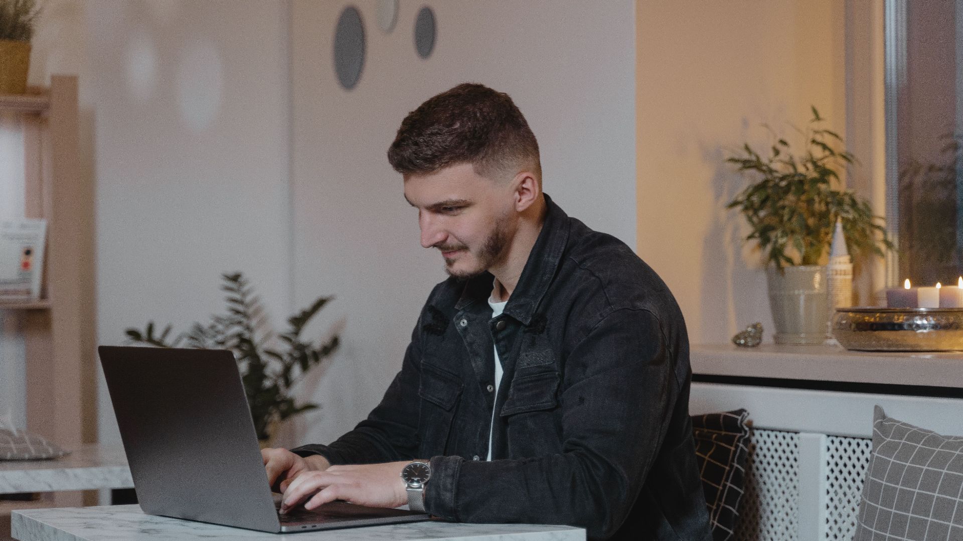 Caucasian man working on a laptop at a cozy indoor setting, surrounded by plants and candles.