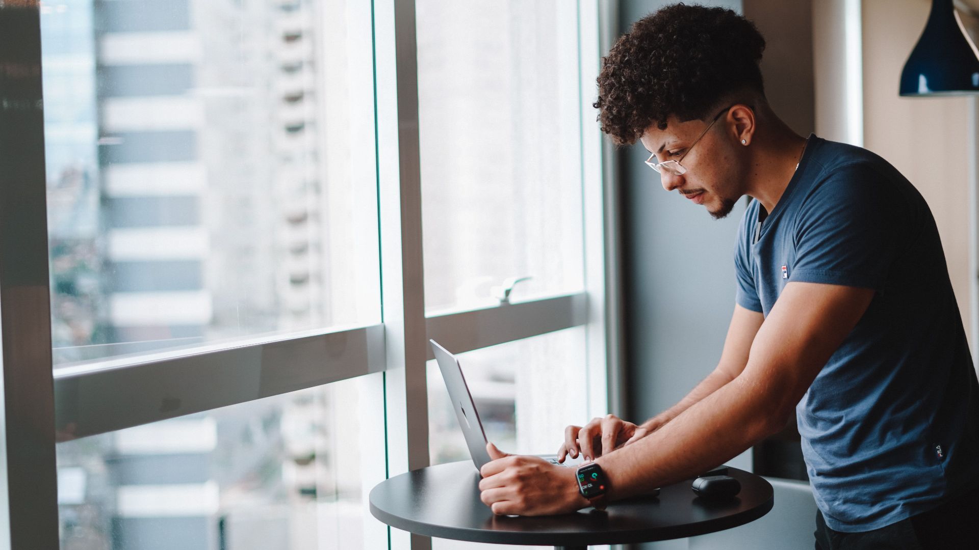 Young man using laptop standing by window in modern office setting.