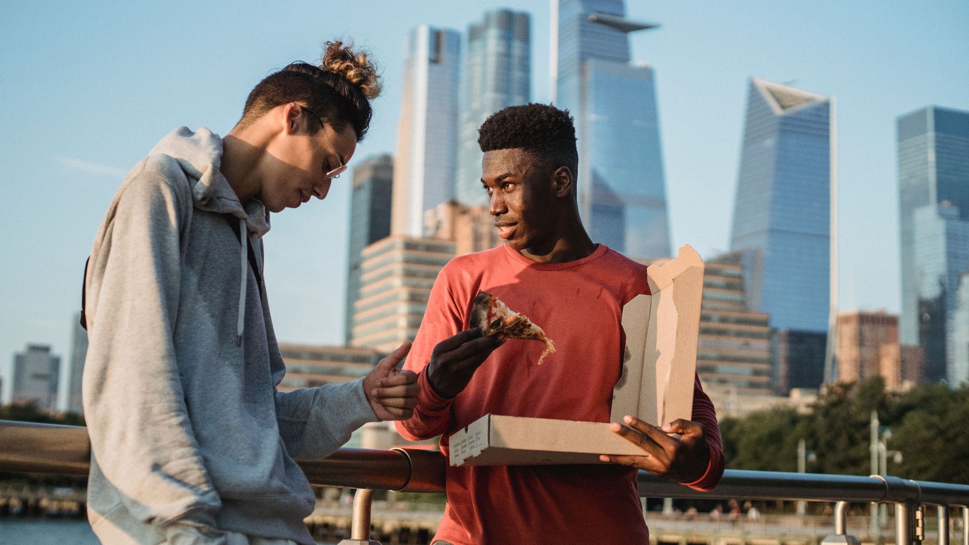 Content black man offering yummy pizza to male friend while spending summer day on modern city embankment