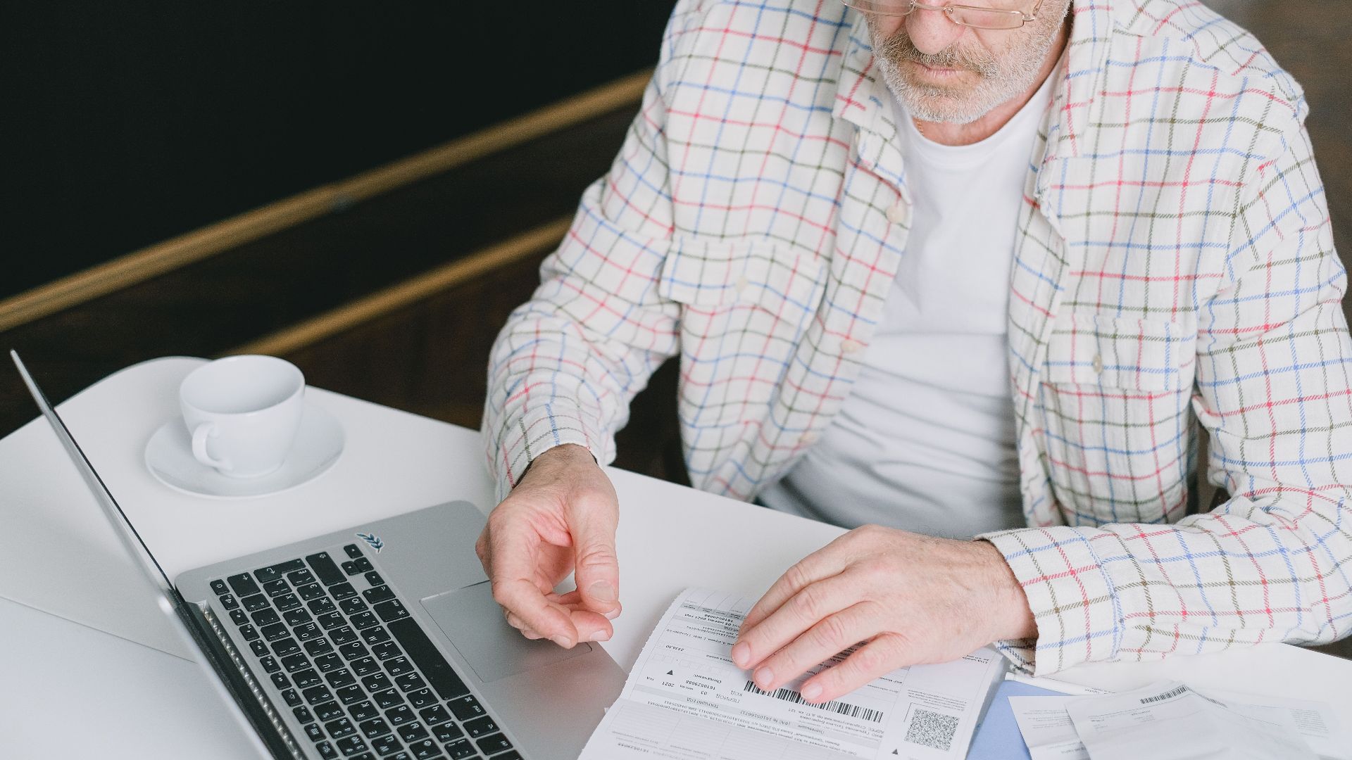Elderly man with eyeglasses reviewing documents at a laptop. Indoor setting with natural light.
