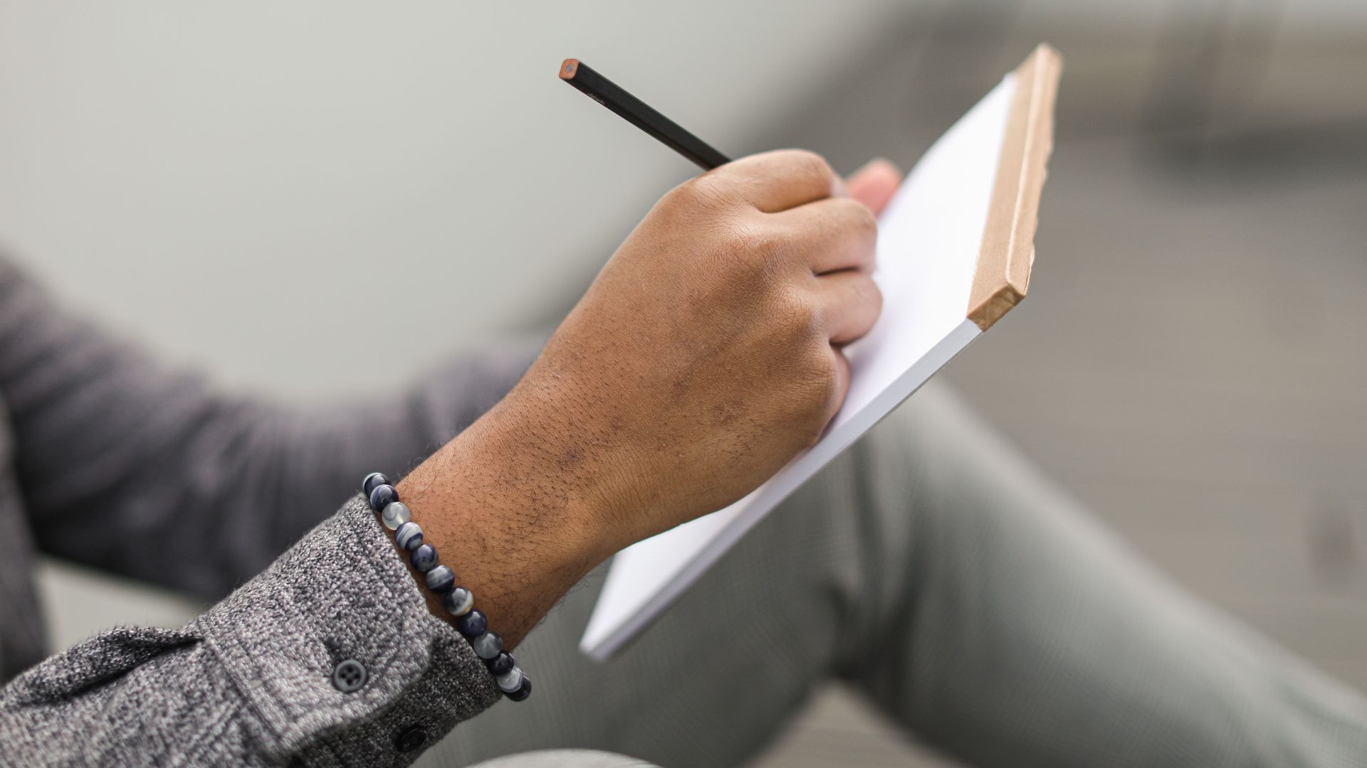 An adult hand writes in a notebook with a pencil, wearing a beaded bracelet and gray plaid sleeves.