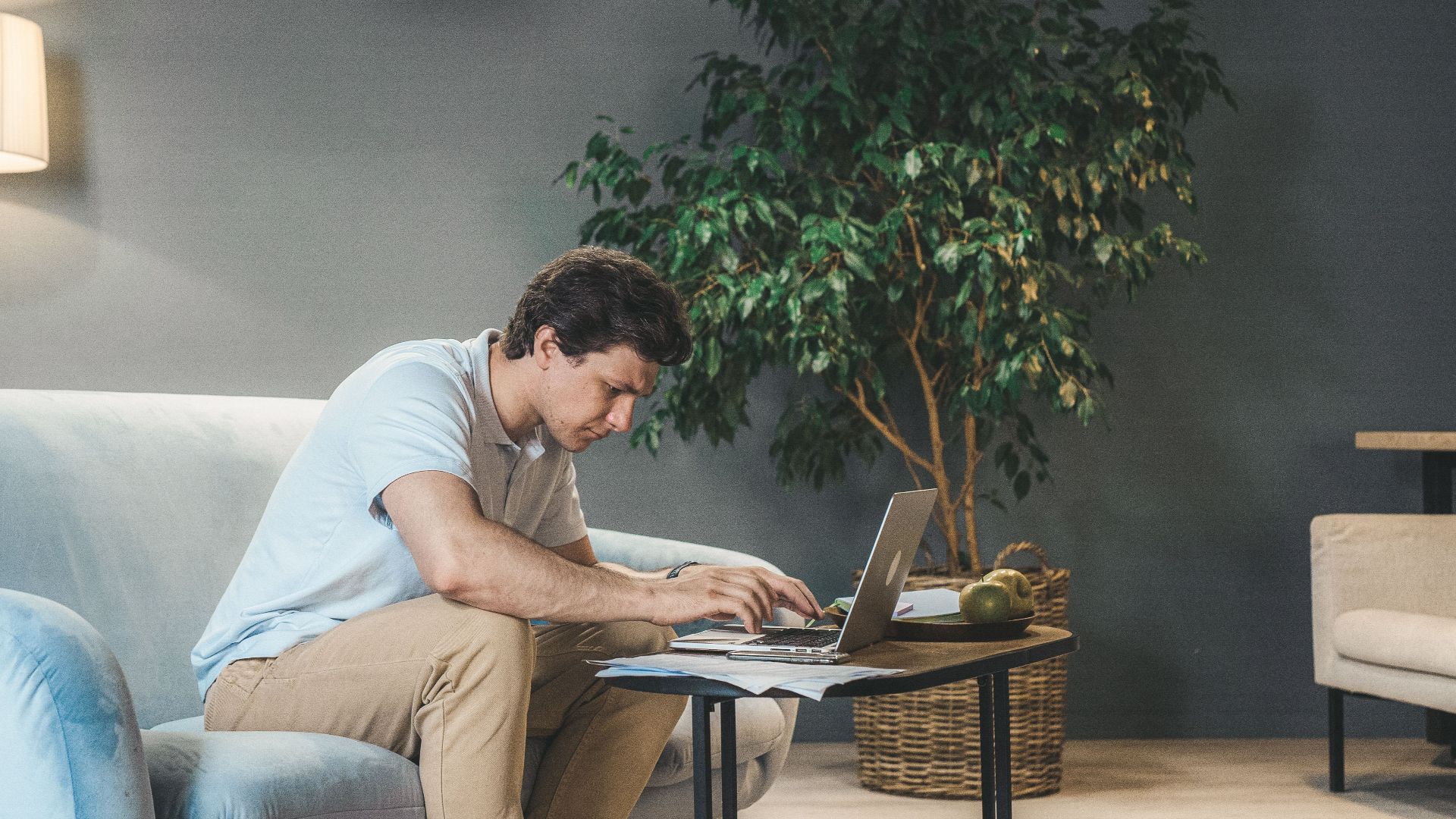 A man working on a laptop in a cozy living room setting with a sofa and a wooden table.